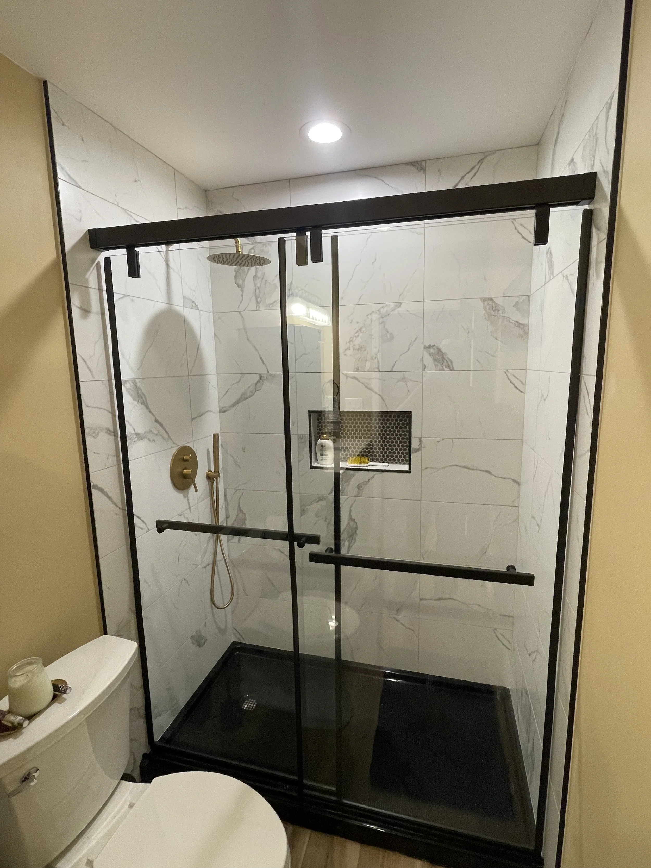 A modern walk-in shower with black metal framing, white marble tiles, a rain showerhead, and a built-in shelf, located in a bathroom with beige walls and a white toilet.