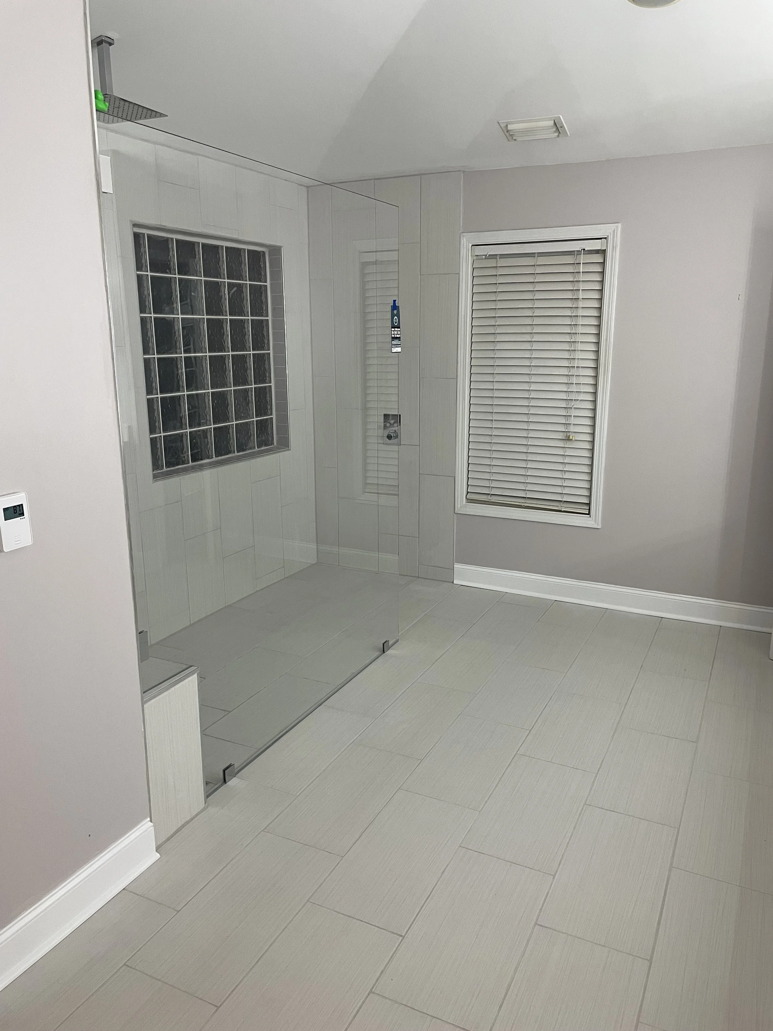 A spacious bathroom with a glass shower enclosure, a window with closed blinds, and light-colored tiled flooring and walls.