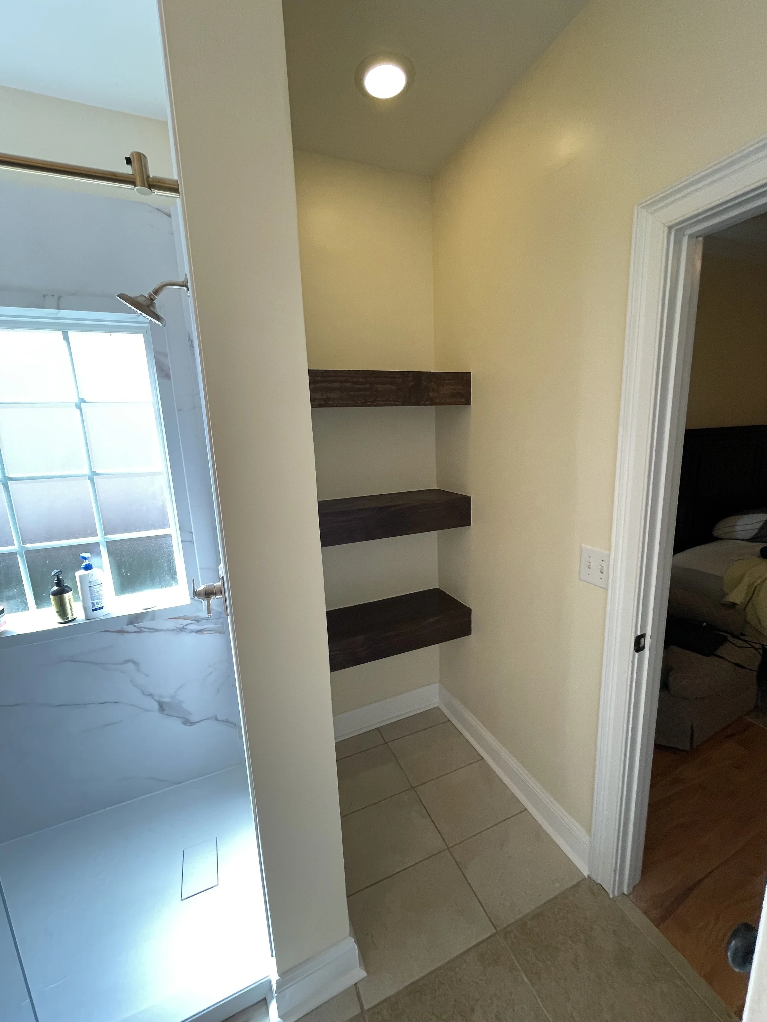 Small corner shelves made of dark wood attached to a beige wall, near a doorway leading to a bedroom with a bed and wooden furniture.