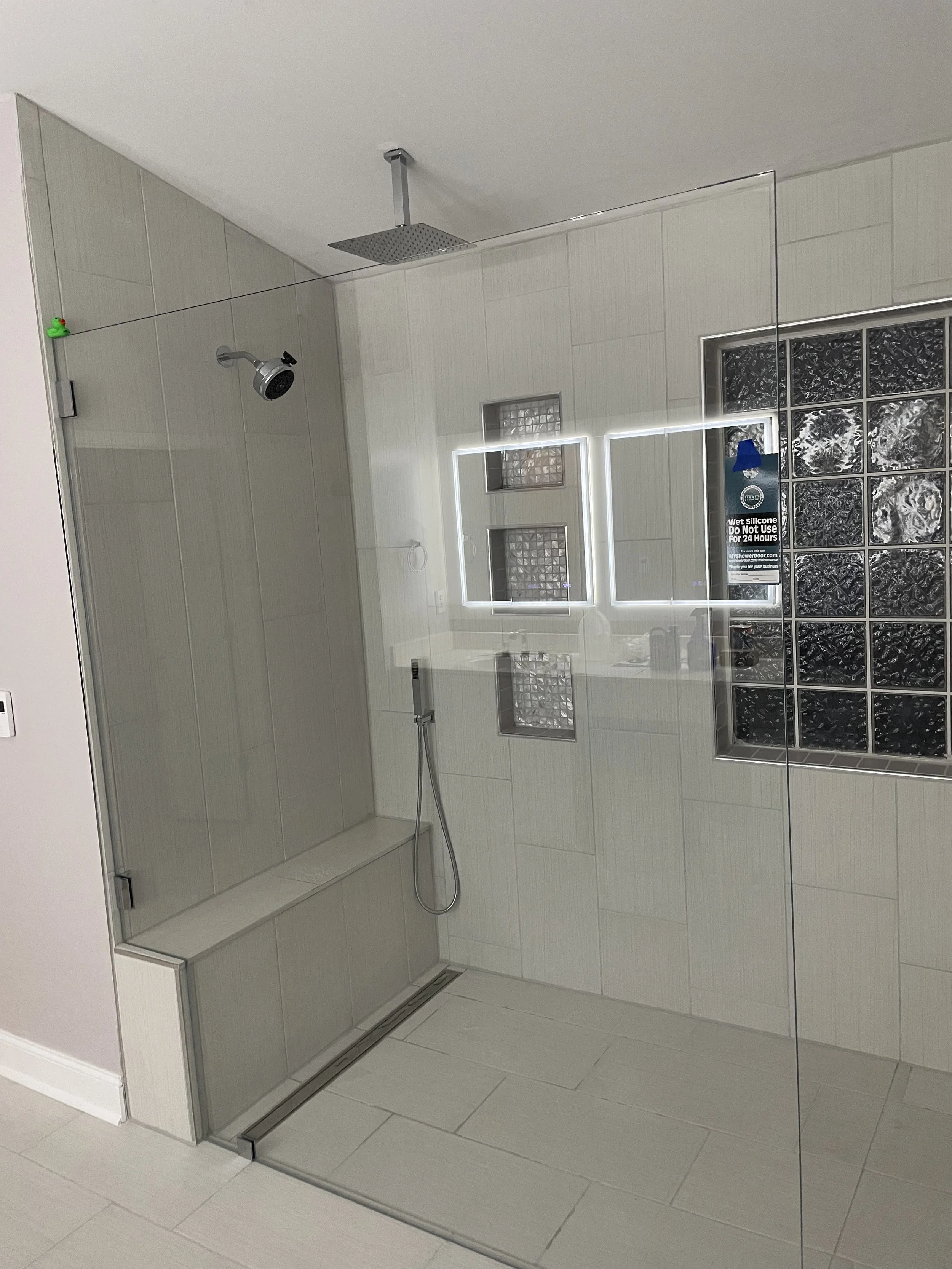 Modern walk-in shower with a glass enclosure, rainfall showerhead on the ceiling, a separate handheld shower, and a small textured glass block window, with a black tiled section and signage to prevent silicone use.