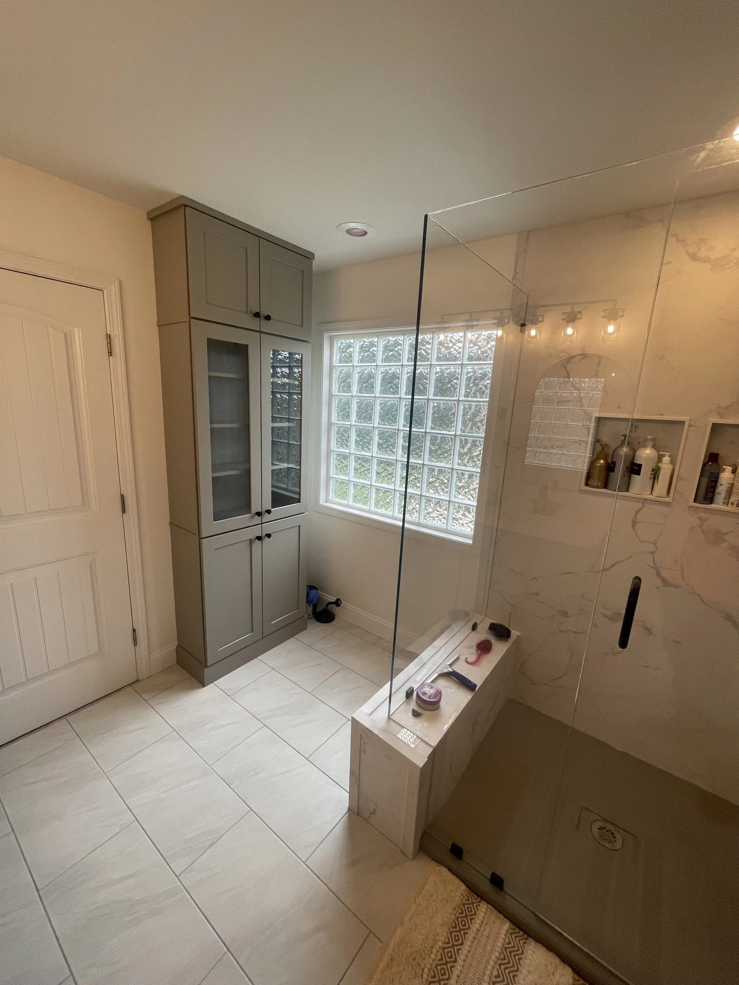 Bathroom with a built-in cabinet, a glass block window, tiled floor, and a shower area with a marble wall. Items on a small ledge include tools and supplies.