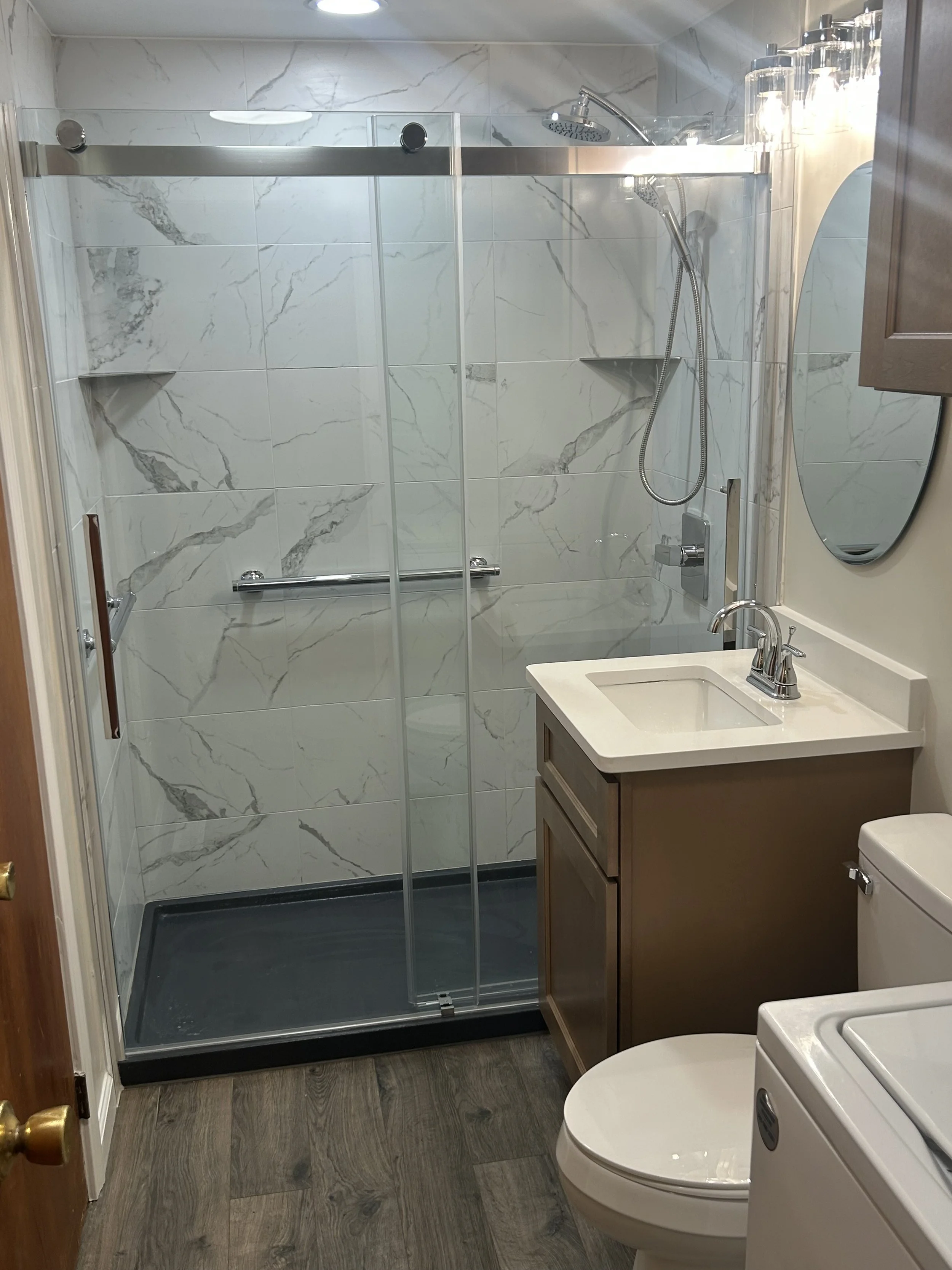 A small bathroom with a walk-in shower with a glass door, a white sink with a wooden cabinet, and a white toilet. The shower has marble-patterned tiles, and there is a mirror on the wall next to the sink. The bathroom floor is wooden.