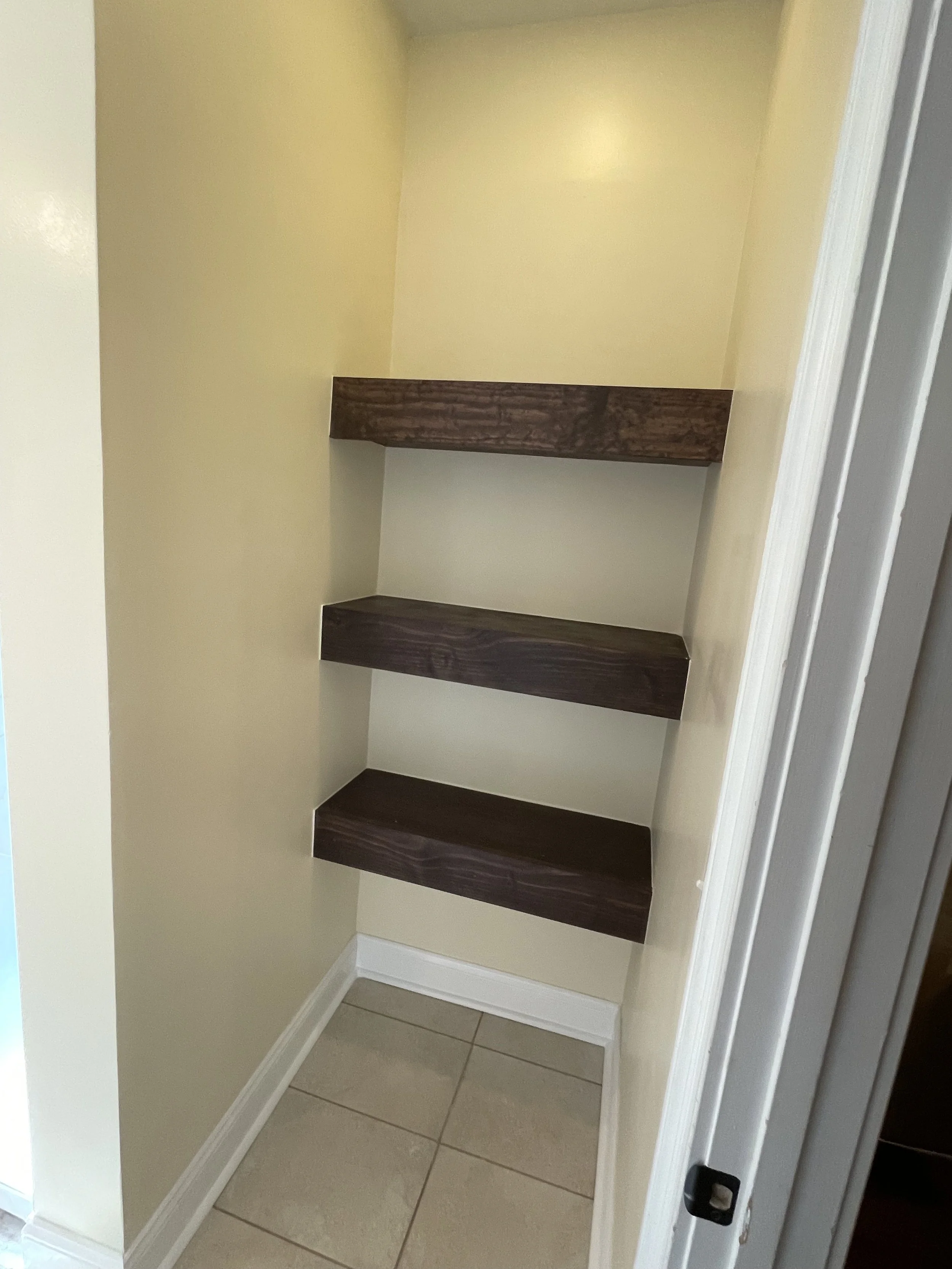 Empty closet with beige walls, four dark wooden shelves, and beige tiled floor.