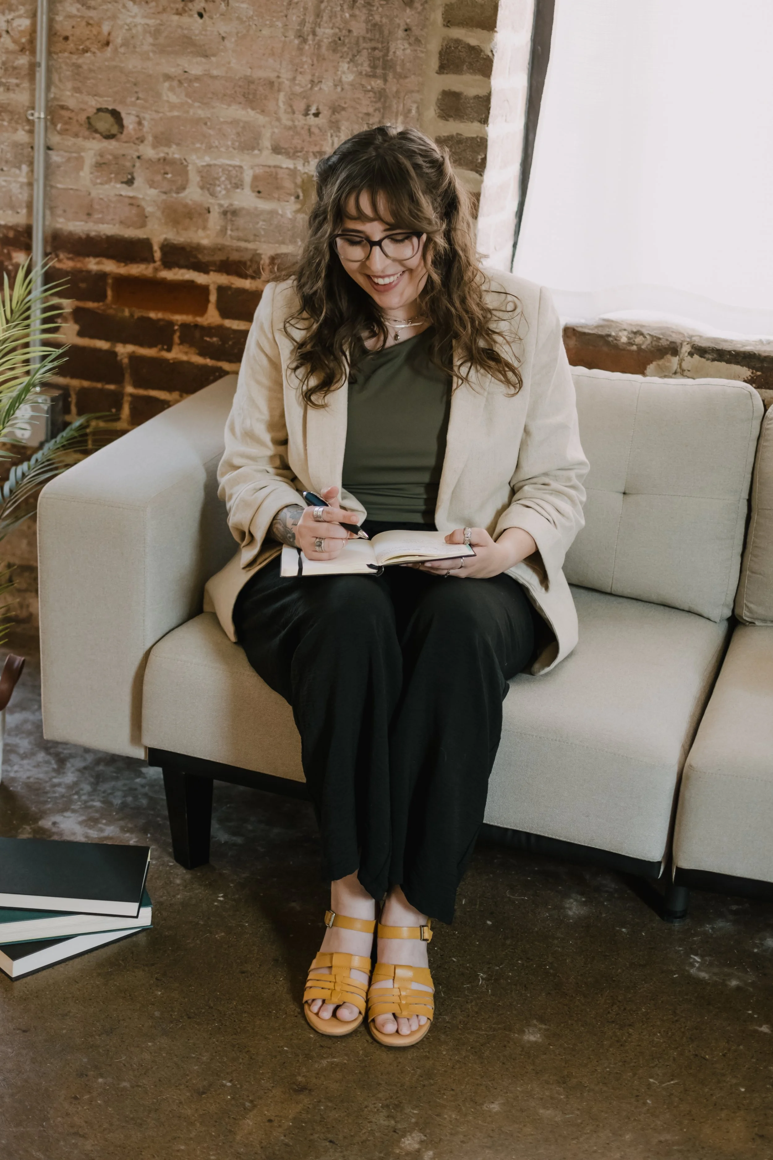 A woman sitting on a beige sofa, writing in a notebook. She has long wavy hair, wears glasses, a beige blazer, a green top, black pants, and yellow sandals. There is a brick wall behind her and a large window with white curtains to her right. A plant is visible on the left side, and some books are on the floor.
