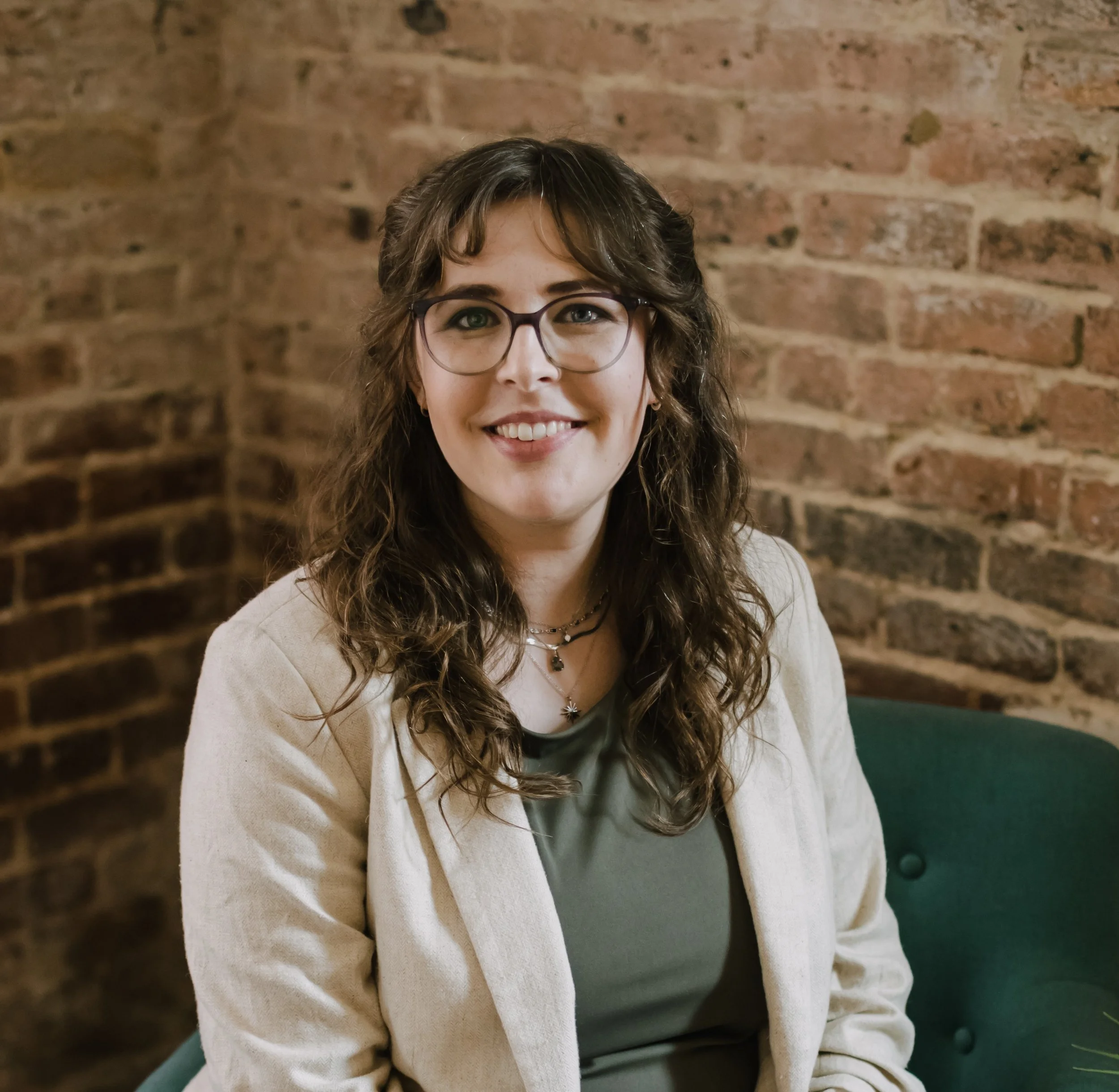 A woman with light skin, long curly brown hair, wearing glasses, a black top, and a beige cardigan, smiling in front of a brick wall.