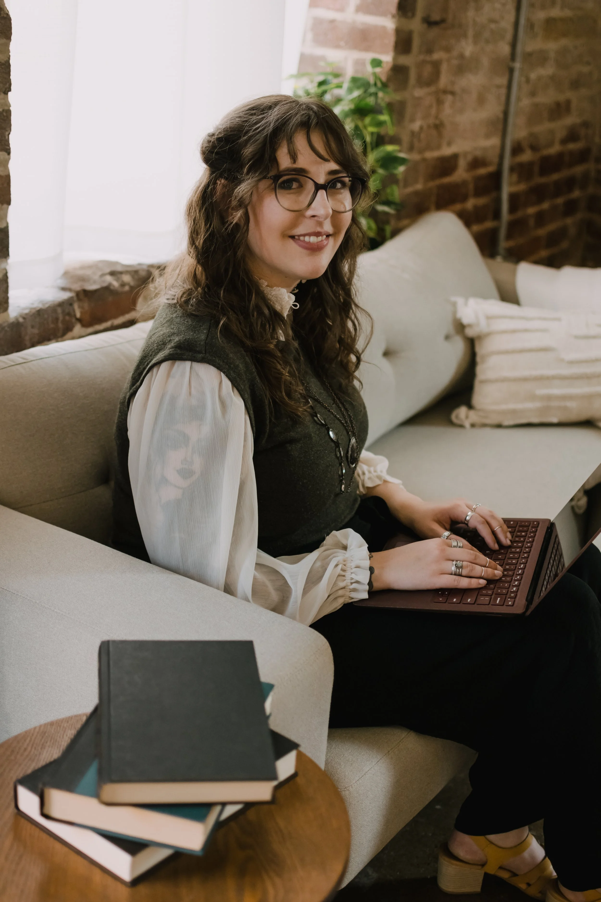 A woman with long curly hair, glasses, wearing a blouse and vest, sitting on a beige sofa with a laptop on her lap, in a room with brick walls and a window with sheer curtains, smiling at the camera.