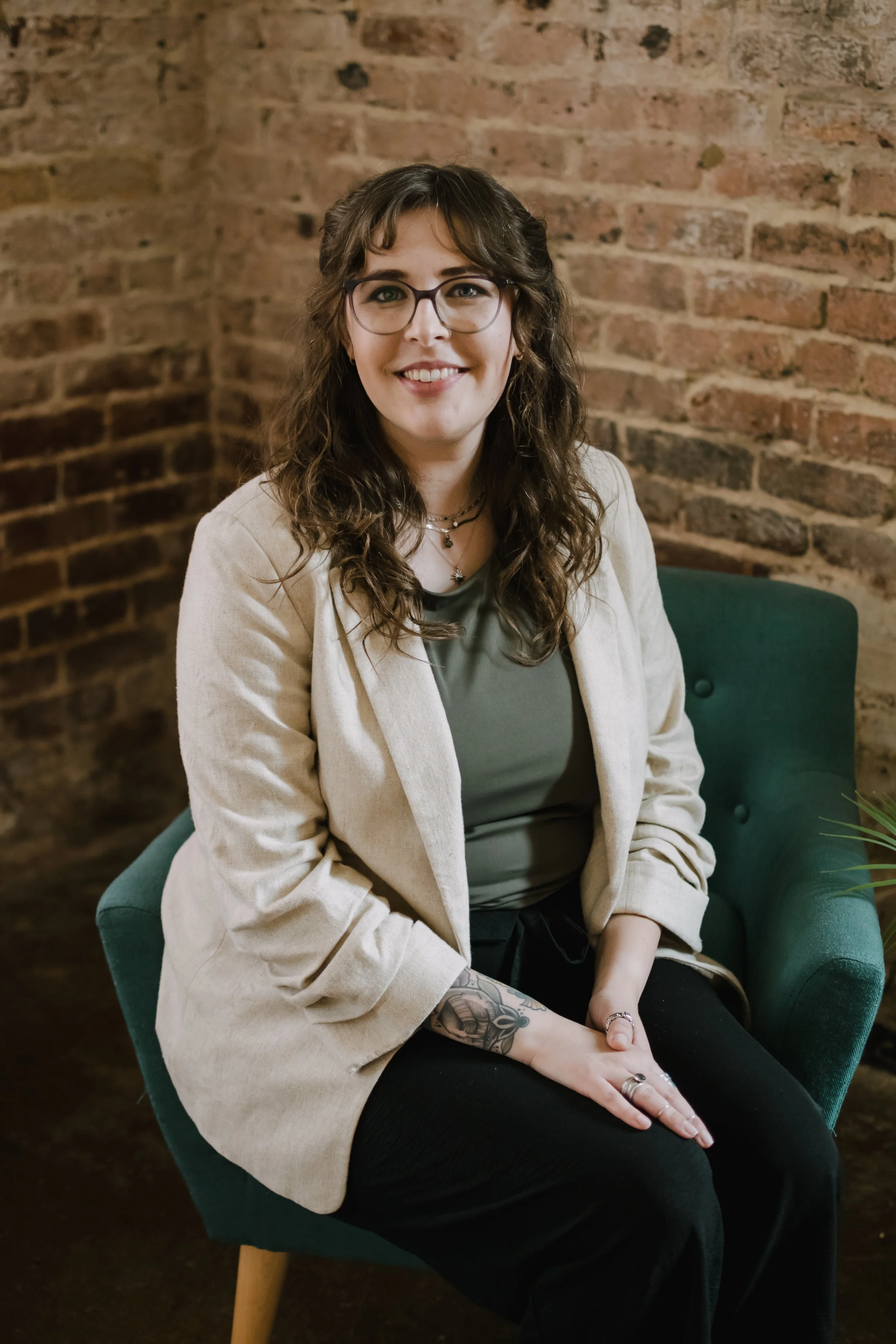 A woman with brown curly hair, glasses, and tattoos on her arm sitting on a green chair in front of an exposed brick wall, smiling and wearing a beige blazer, gray top, and black pants.