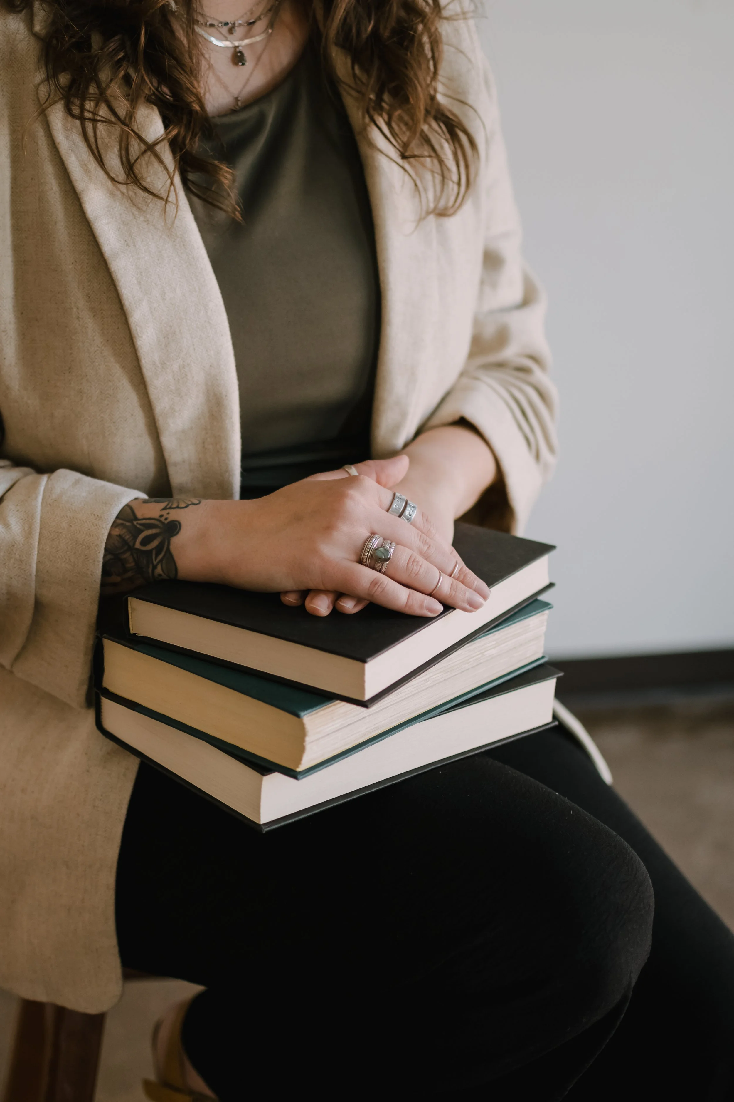 A person wearing a beige blazer and black pants is sitting and holding a stack of books. The person has a tattoo on their forearm, multiple rings on their fingers, and is wearing layered necklaces. The focus is on their hands and the books, with the person's face not visible.
