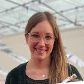 A smiling woman with glasses and long blonde hair, wearing a dark sweater, standing indoors with a modern architectural background.