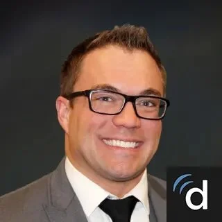 A smiling man with short brown hair, glasses, wearing a gray suit, white shirt, and black tie, against a dark background.