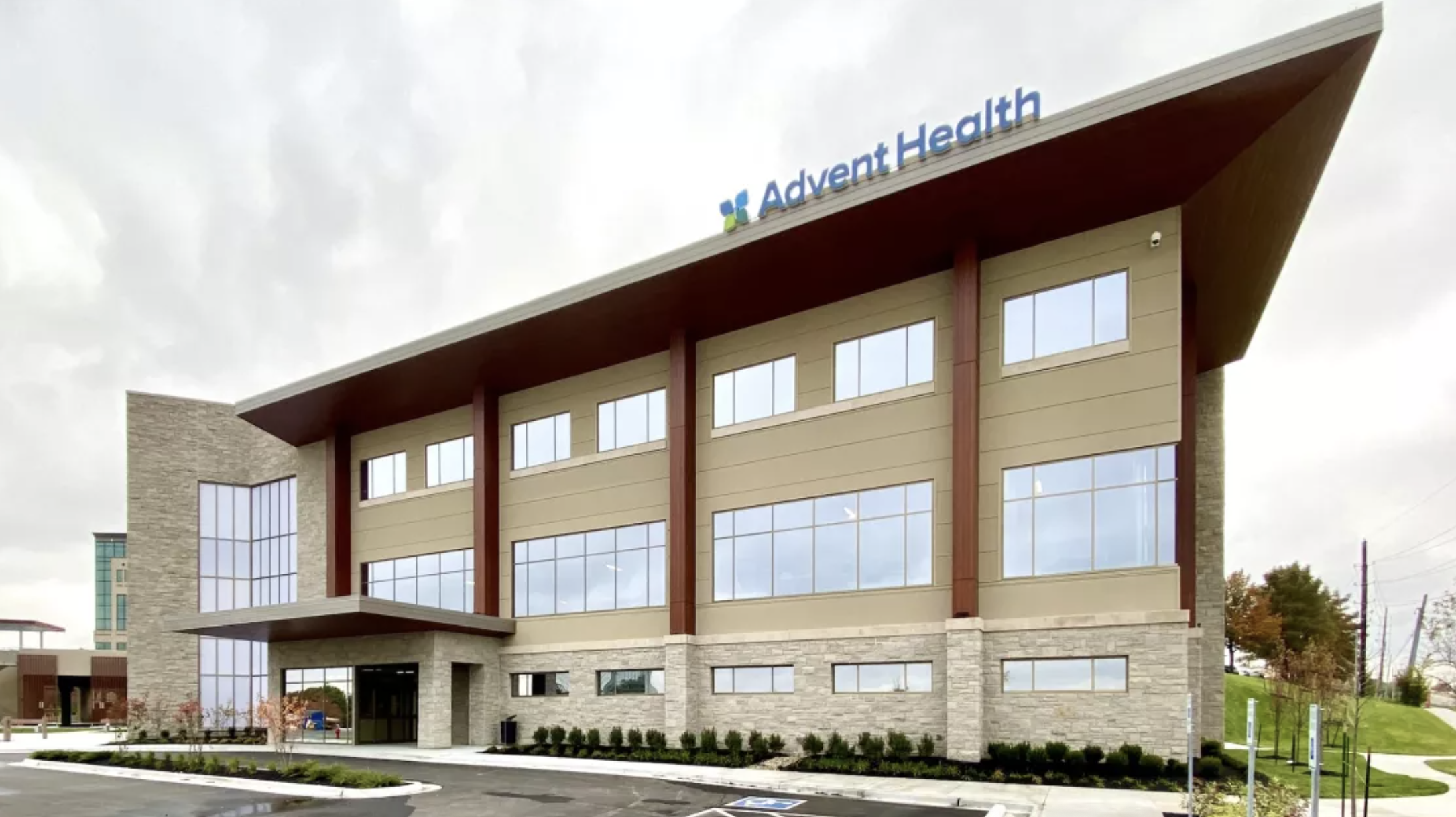 Exterior of a modern building labeled Advent Health, with large windows, stone and wood accents, and a landscaped parking lot.