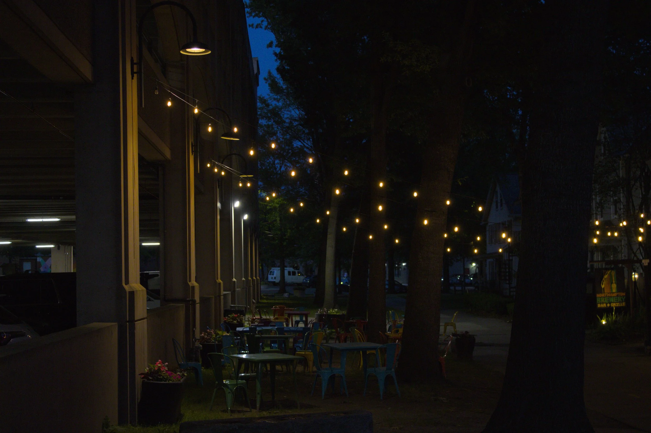 Nighttime outdoor patio with strings of hanging yellow lights, trees, and empty colorful chairs and tables.