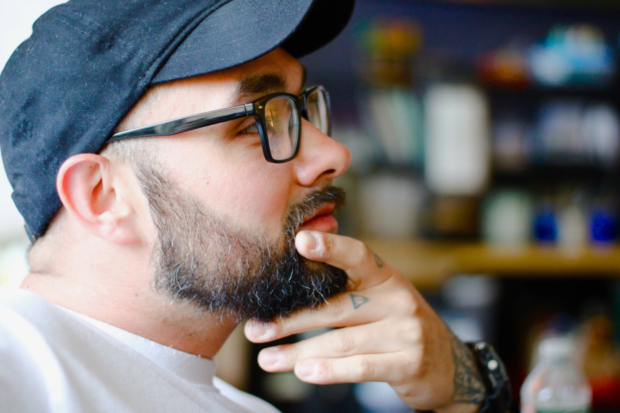 Side profile of a man with glasses and a beard, wearing a black cap and a white shirt, touching his chin with his hand in a thoughtful pose, with blurred shelves of bottles and containers in the background.