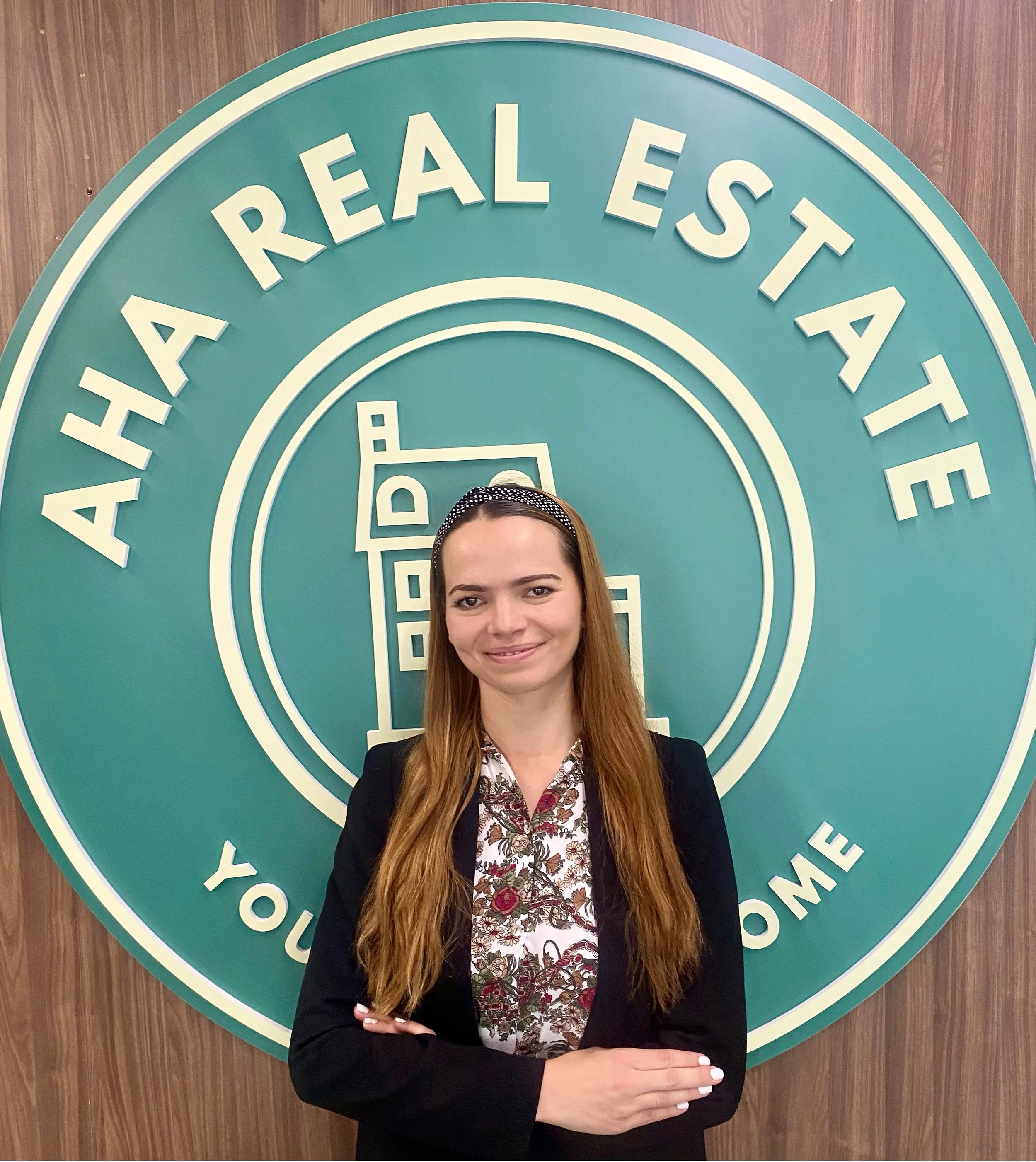 A woman with long hair and a headband, wearing a black blazer and a floral blouse, standing in front of a large circular sign that reads 'Atlanta Real Estate' and 'You're Home'.