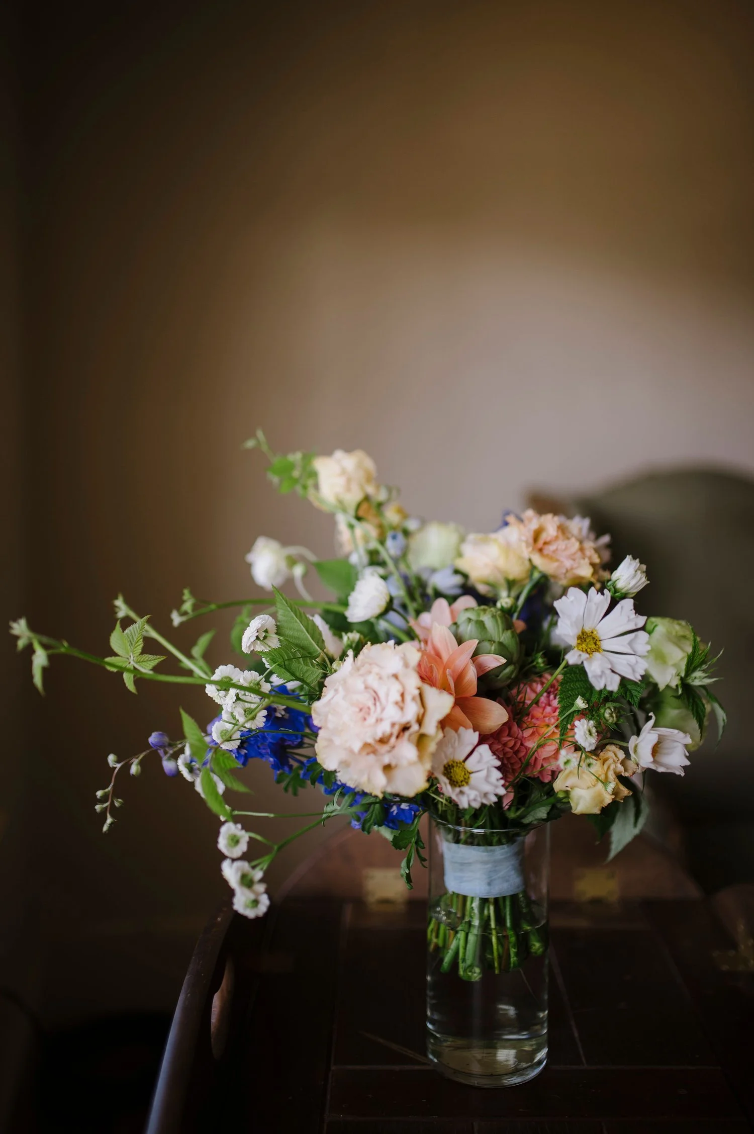 A colorful bouquet of various flowers including carnations, daisies, and other blossoms in a glass vase on a dark wooden table.