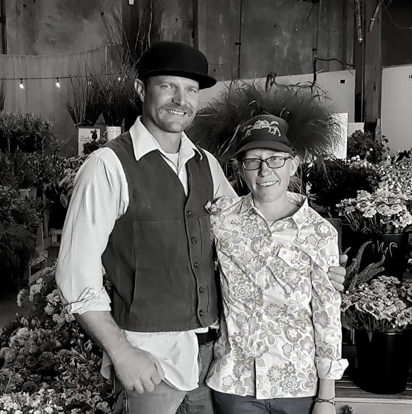 A man and woman standing together in a plant nursery or florist shop, smiling, with various potted flowers and plants in the background.