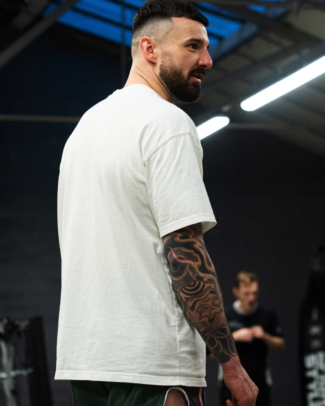 A man with a beard and short dark hair, wearing a white t-shirt, stands in a gym with fitness equipment in the background. He has a tattoo sleeve on his left arm and is looking to the side.