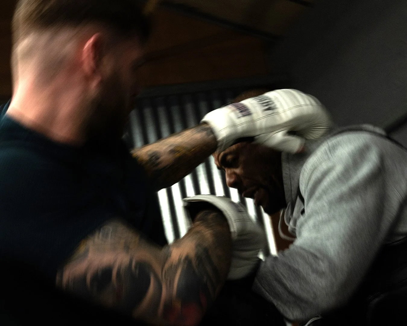 Two men engaged in a physical fight or confrontation, one with tattooed arms and the other wearing a gray hoodie, in an indoor setting with vertical black and white striped background.