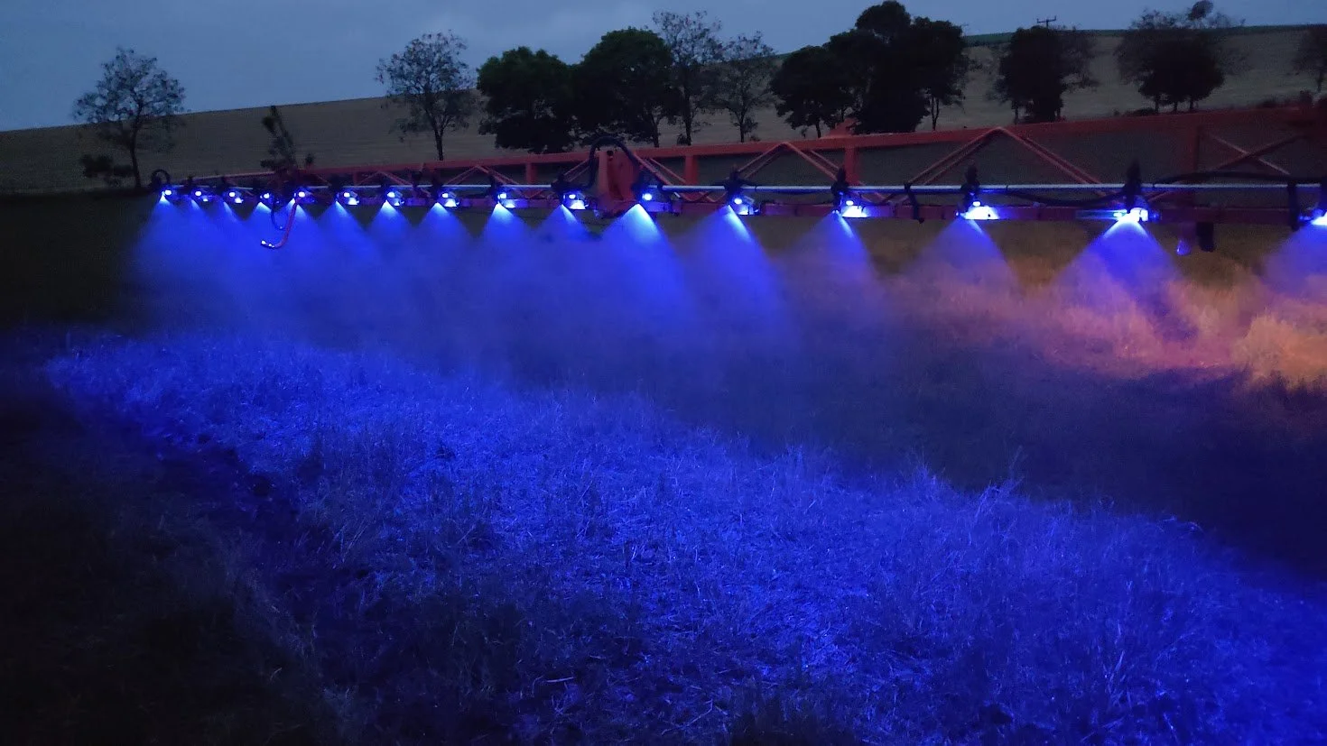 Sistema de irrigação agrícola funcionando em campo com irrigadores acesos, emitindo jatos de água sob luzes azuis, com árvores e colina ao fundo ao entardecer.