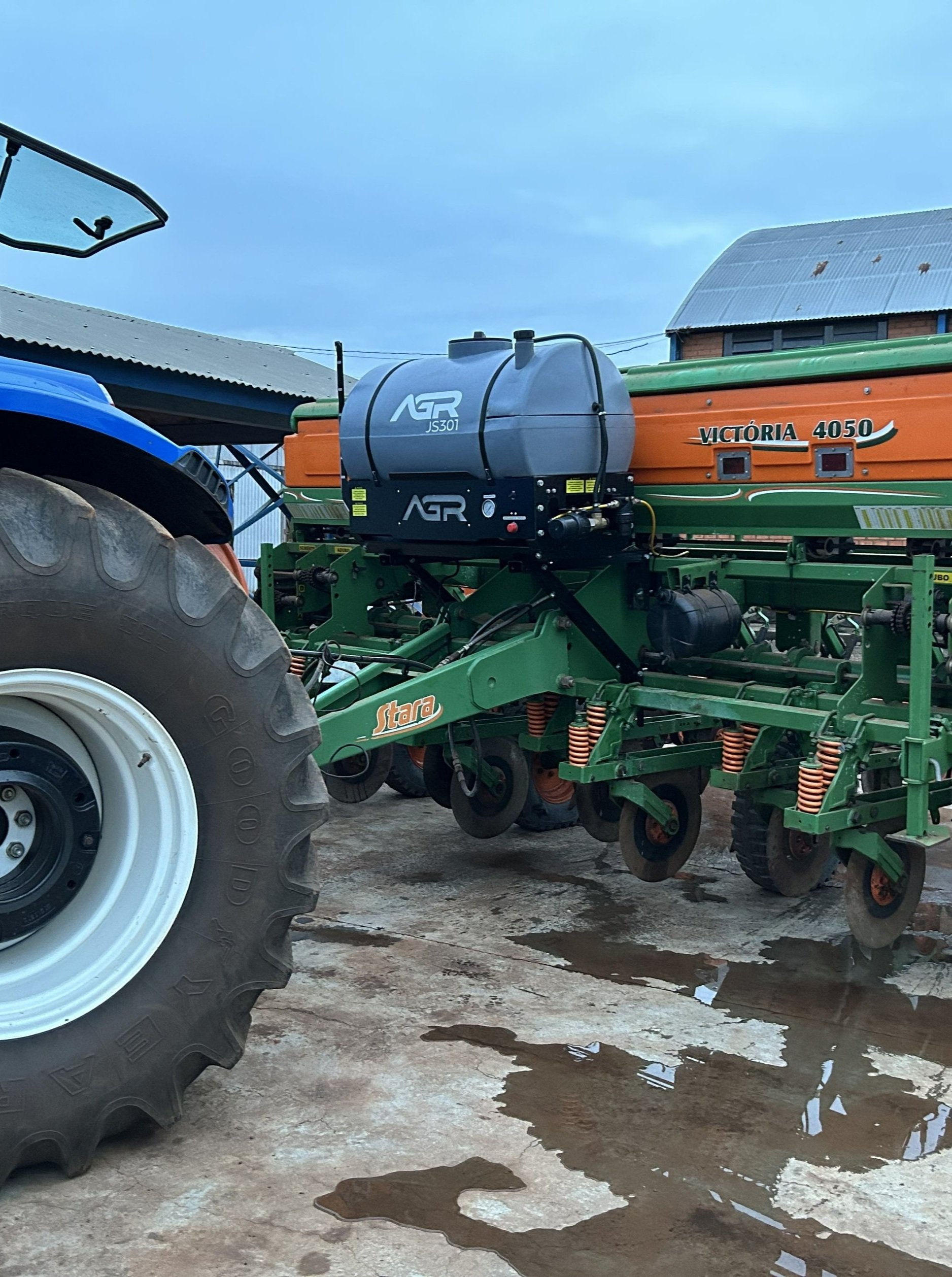 Máquina agrícola estacionada ao lado de um trator, com piso encharcado e céu nublado ao fundo.