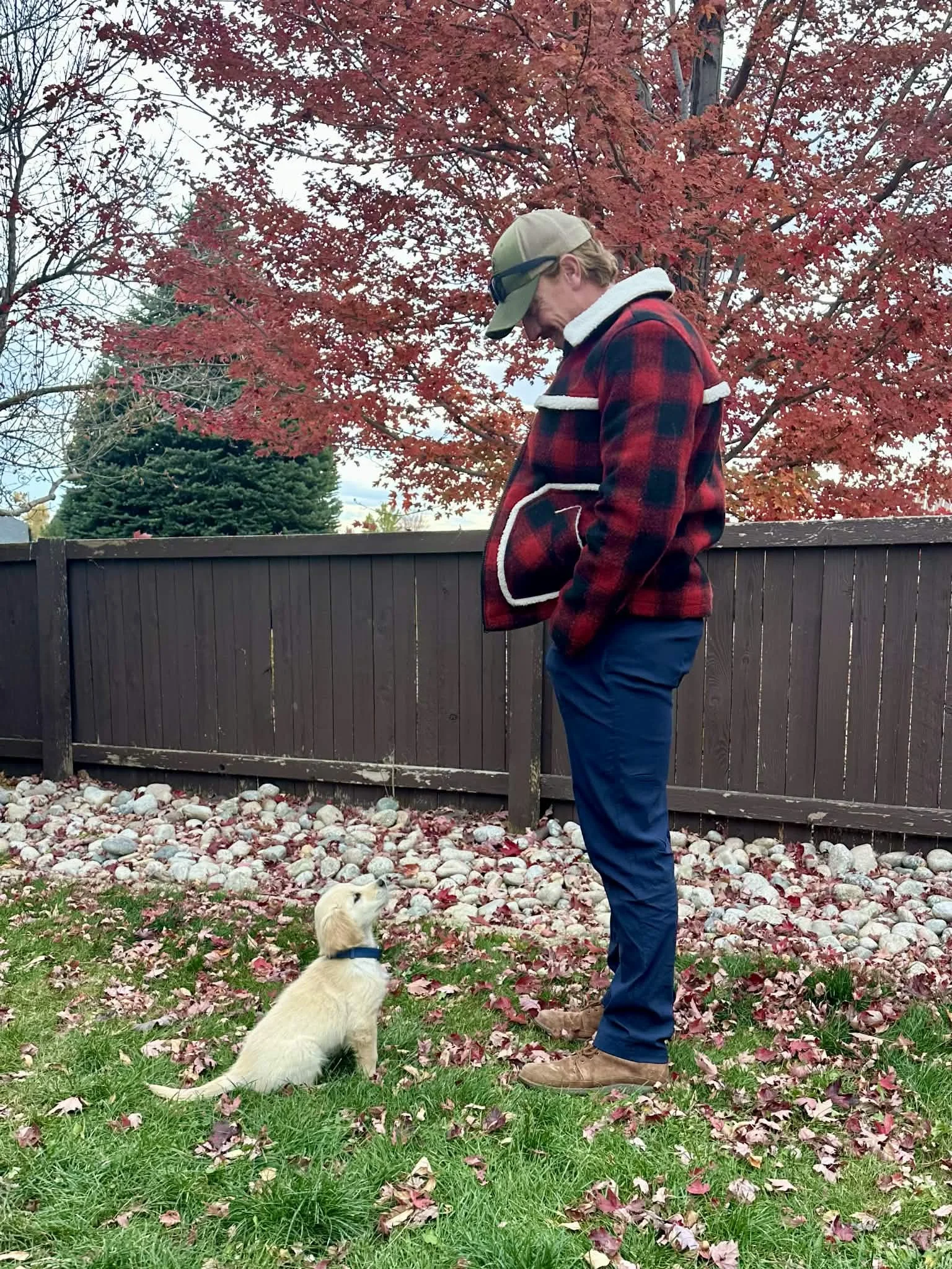 A man wearing a red and black plaid jacket, beige cap, and sunglasses stands outdoors in front of a red autumn tree, with a small cream-colored dog sitting on the grass at his feet, surrounded by fallen pink and red leaves.