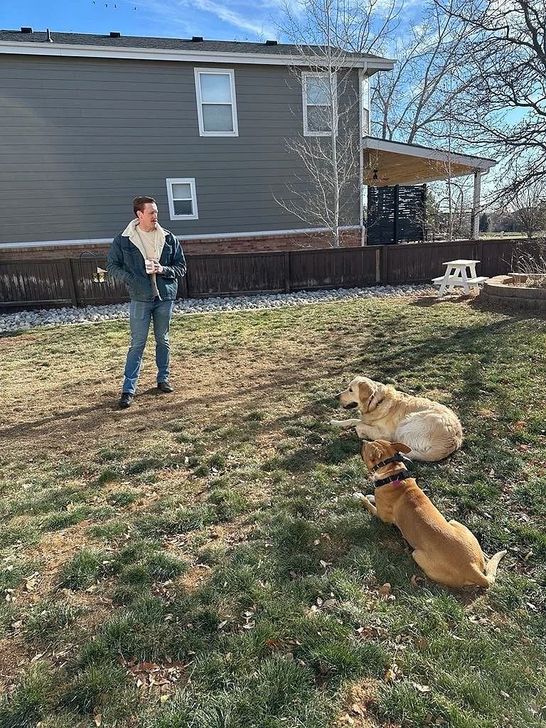 A man standing outside in a yard with two dogs lying on the grass, all facing the man. The yard has a house in the background, a wooden fence, and a small white picnic table.