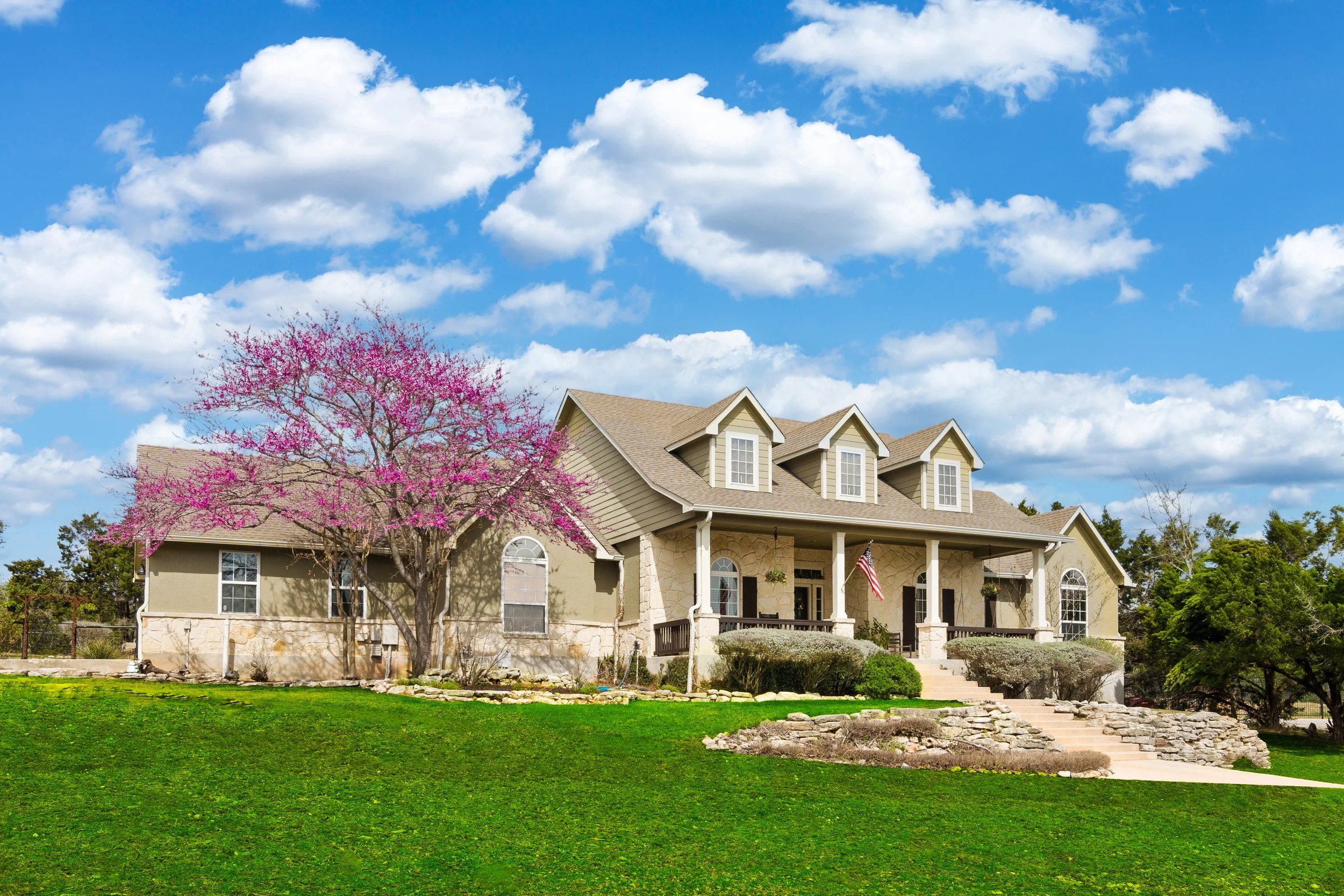 A large two-story house with beige and stone exterior, multiple windows, and a front porch with white columns, surrounded by a well-maintained lawn and trees, including a pink flowering tree, under a partly cloudy blue sky.