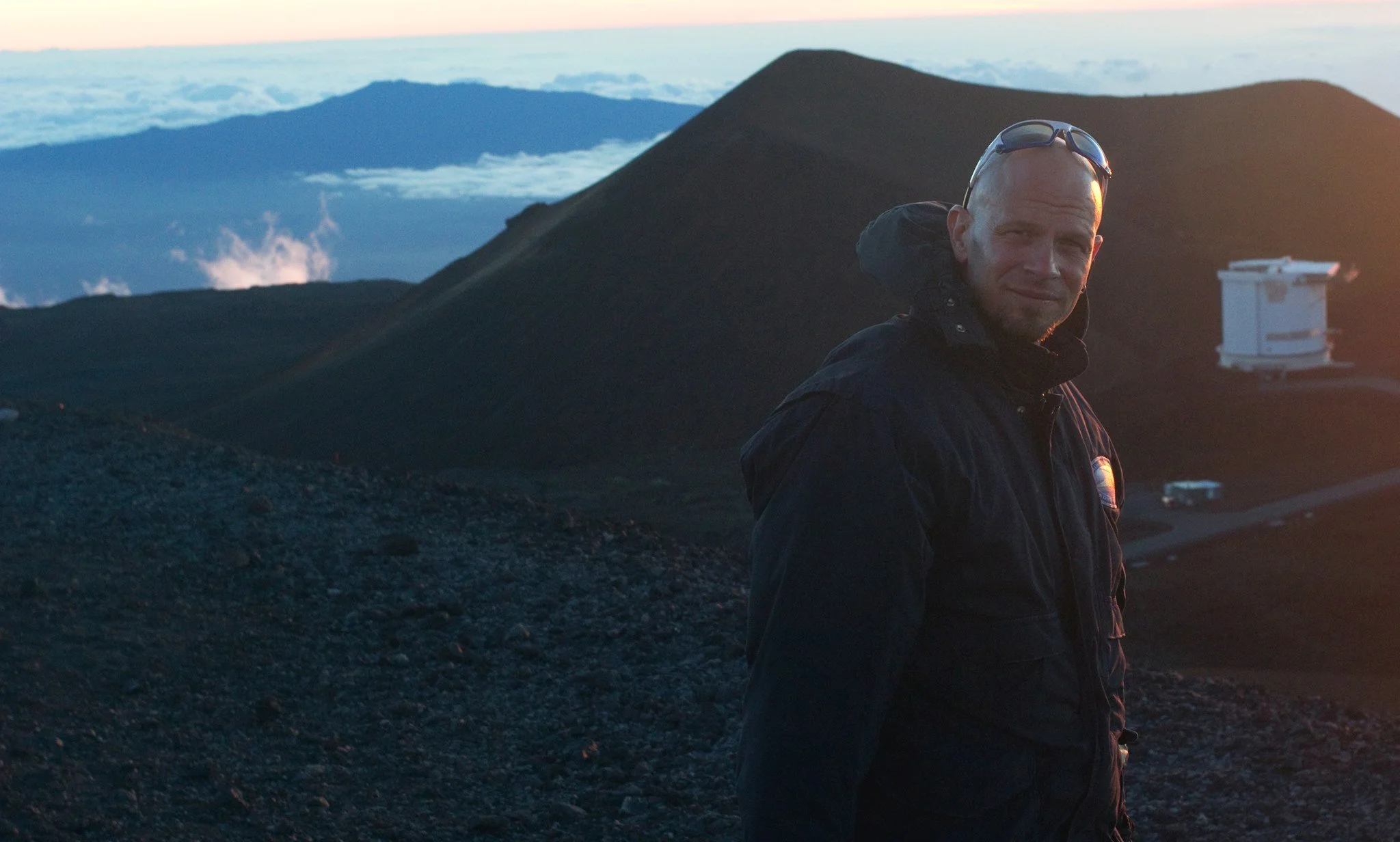 A man with sunglasses on his head wearing a dark jacket, standing on a mountain at sunset, with mountains and clouds in the background.
