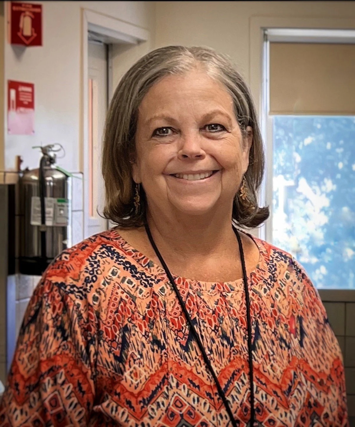 A smiling middle-aged woman with shoulder-length gray hair, wearing earrings, a patterned orange, red, and navy shirt, and a black lanyard, standing indoors next to a window.