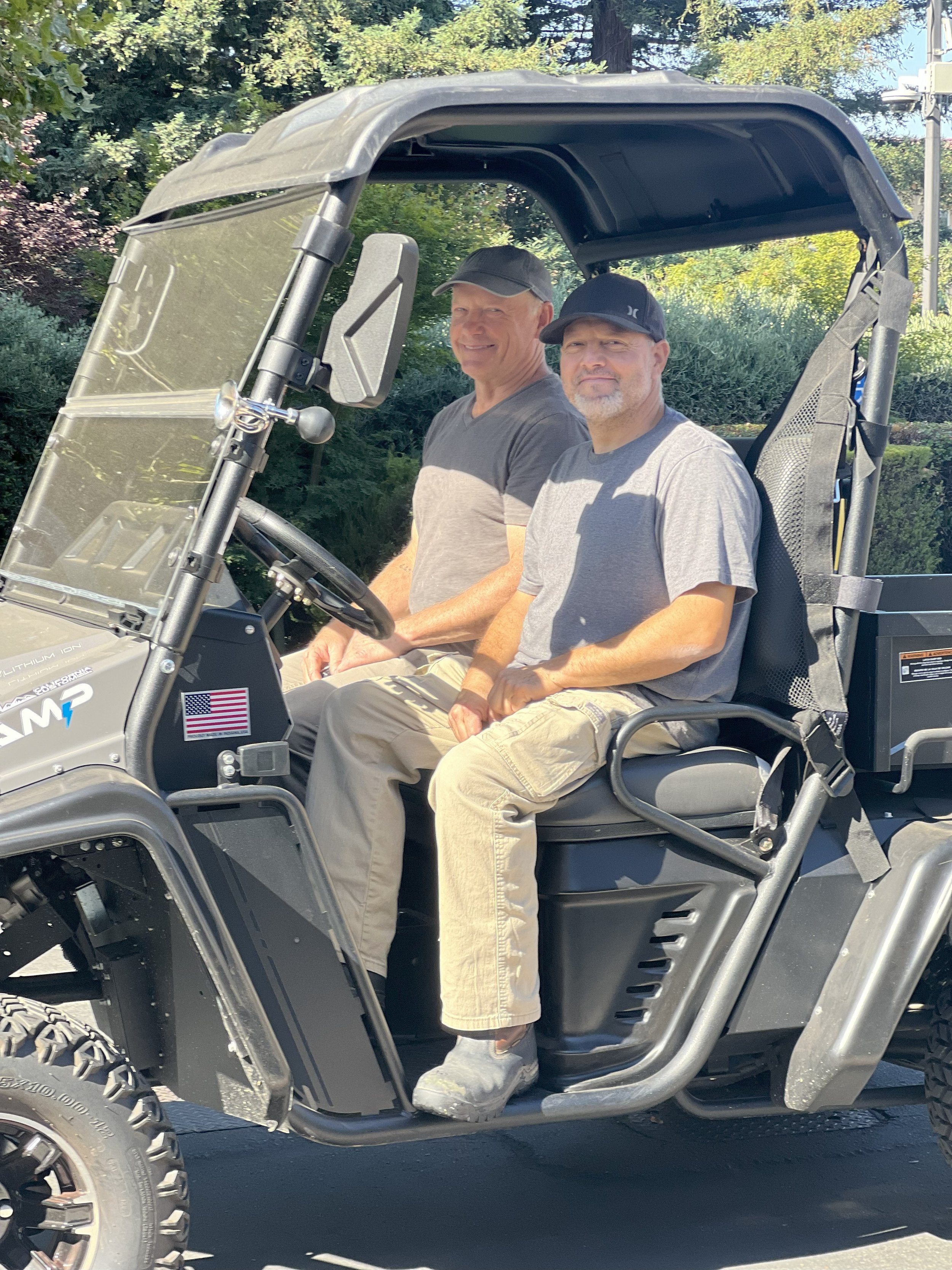 Two men sitting in a utility vehicle, smiling, with trees and greenery in the background on a sunny day.