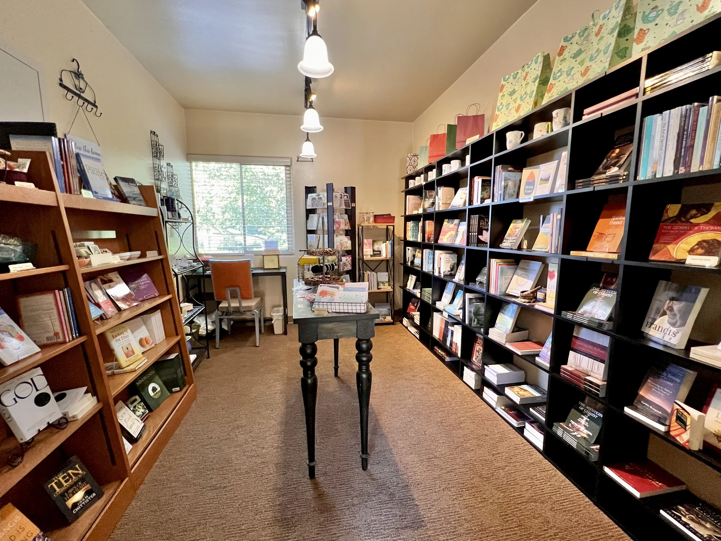 Interior of a small bookstore with bookshelves, a table, and a desk near a window.