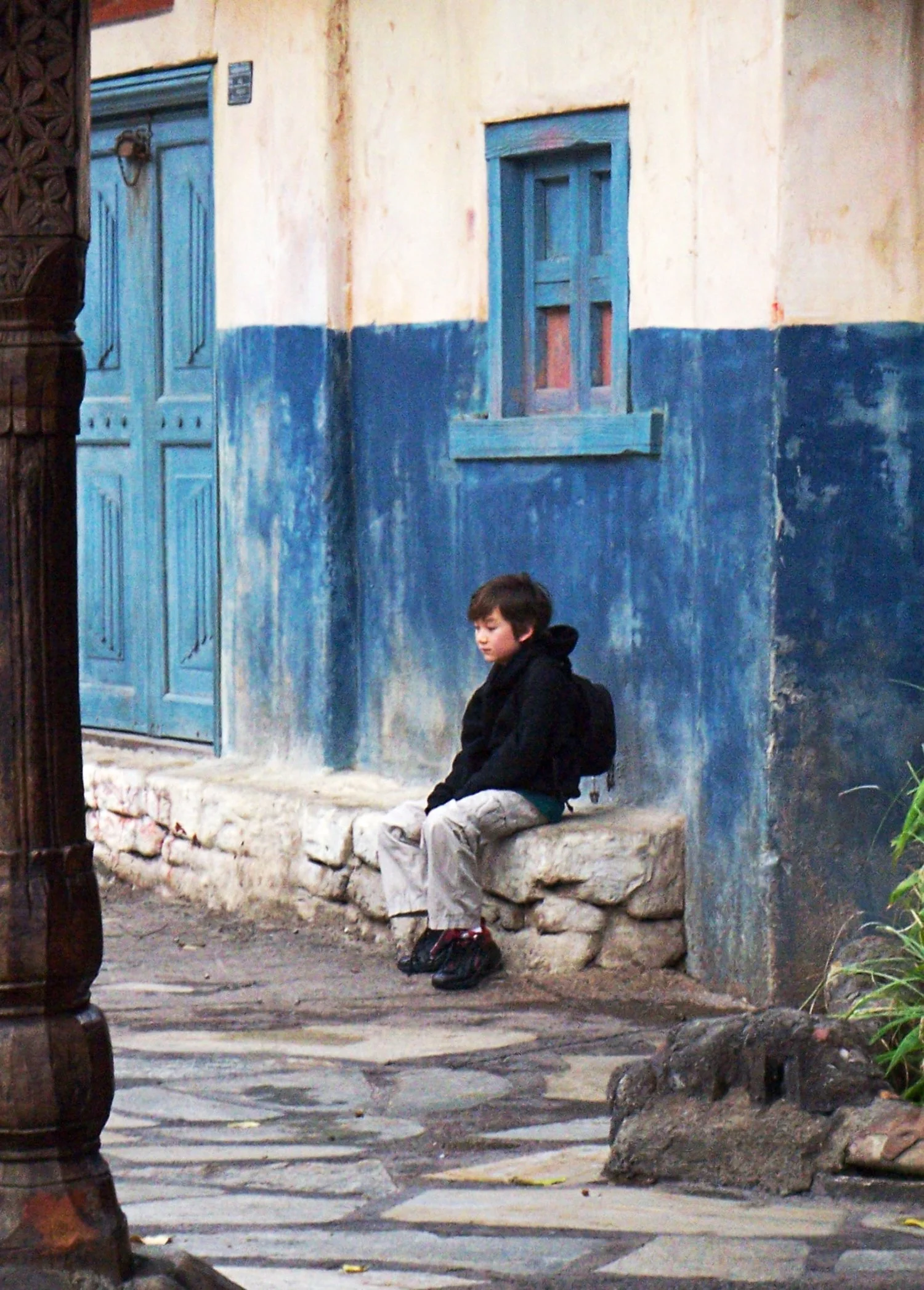 Boy On Wall - Sometimes the quietest moments speak the loudest. One child, one wall, endless stories untold. 
A young boy wearing a black hoodie and light-colored pants sits quietly on a stone ledge against a rustic blue and white wall with a wooden
