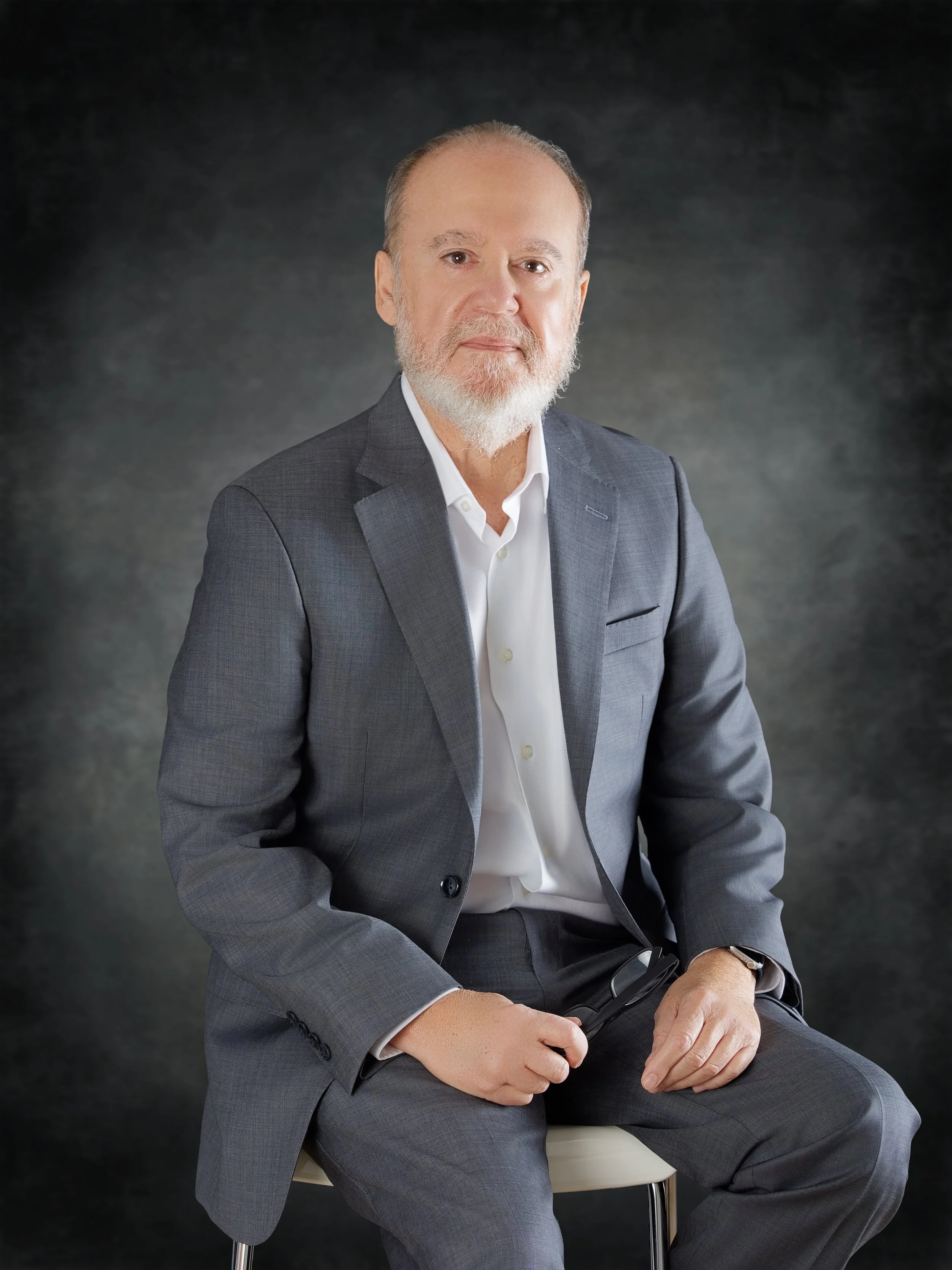 A mature man with a beard wearing a gray suit, white shirt, and glasses sitting on a chair against a dark gray background.