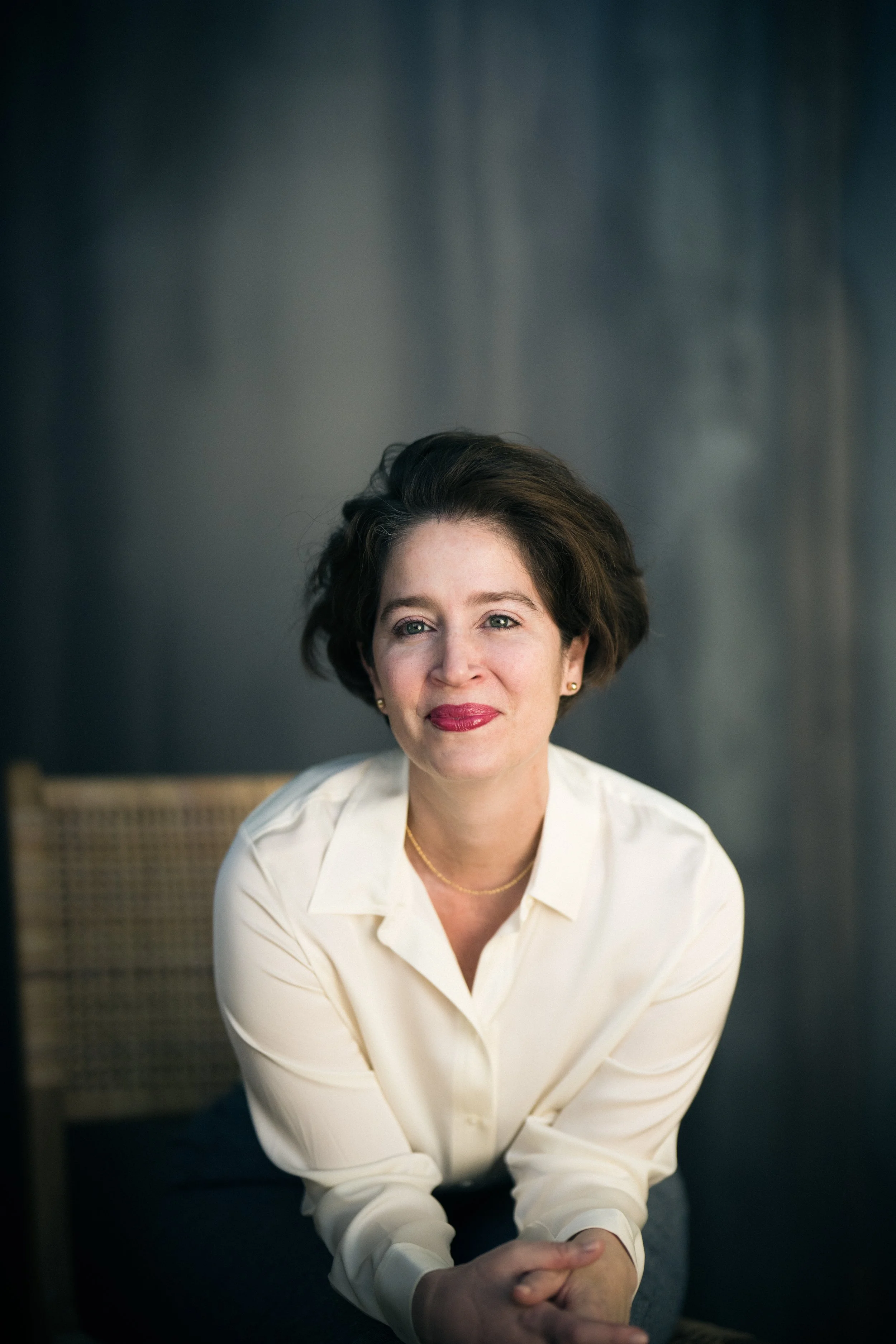 A woman with short dark hair and red lipstick seated in front of a dark background, wearing a cream-colored blouse and smiling softly.