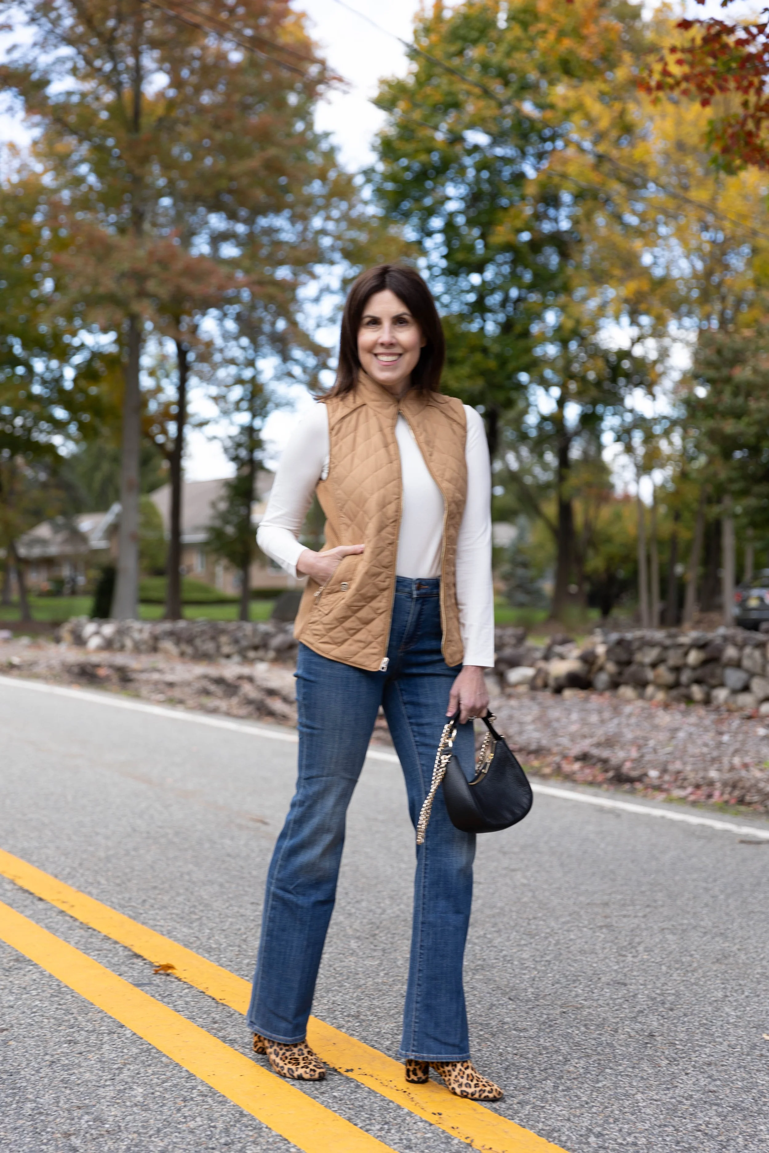 A woman standing on a suburban street during autumn, wearing a tan quilted vest, white long sleeve shirt, blue jeans, leopard print heels, and holding a black purse with chain strap, smiling at the camera.