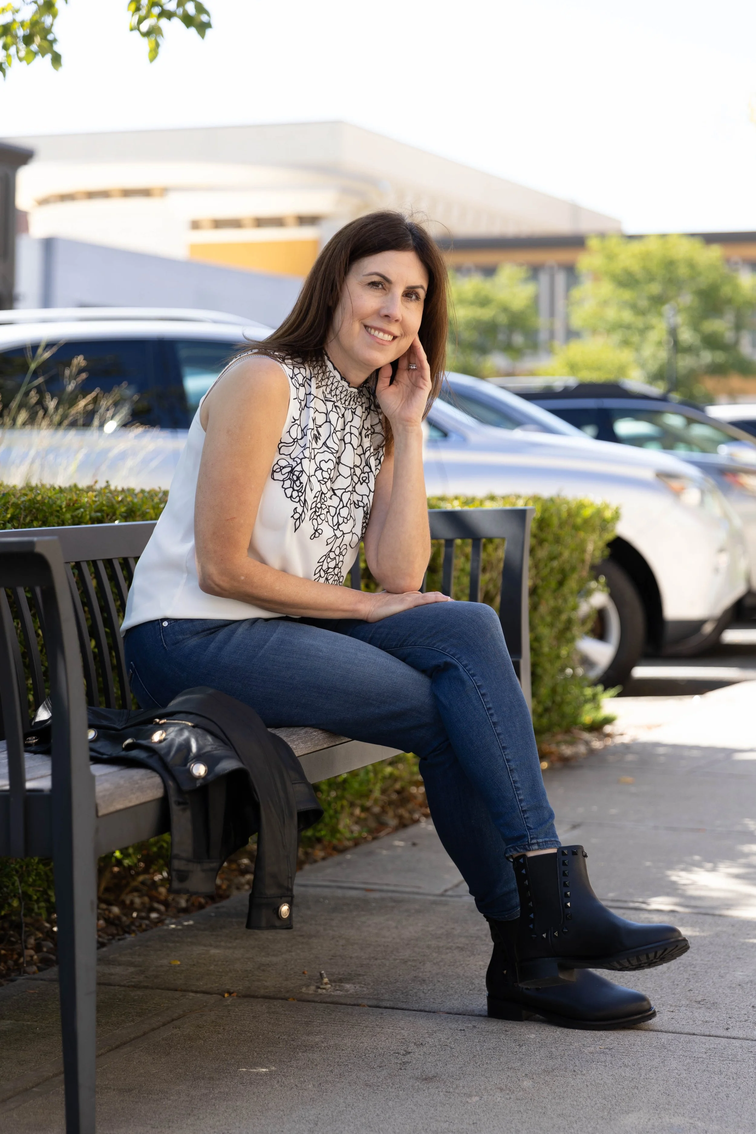 A woman sitting on a park bench with a black leather jacket beside her, smiling and touching her face, with a parking lot and modern building in the background on a sunny day.