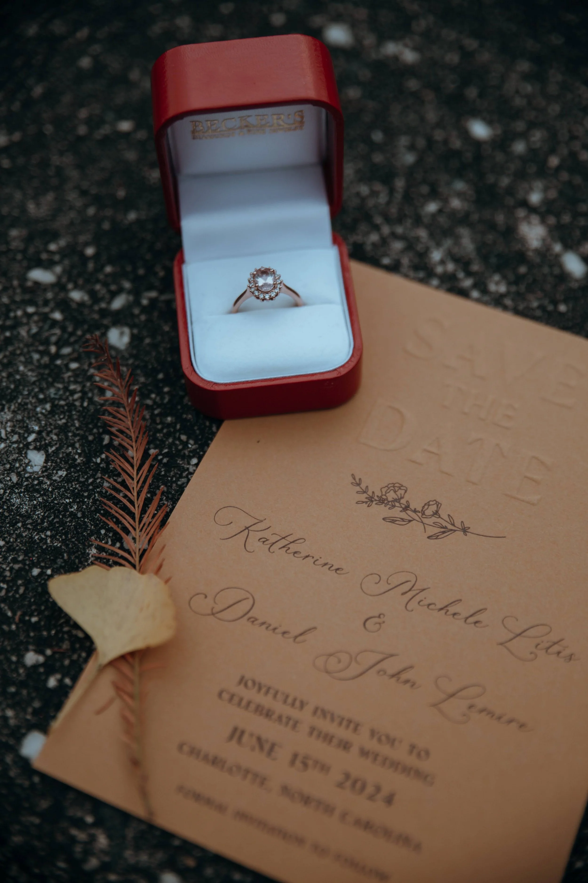 A wedding invitation and a ring in a red box, placed on a dark, textured surface with small white specks, and decorated with a dried flower and leaf.
