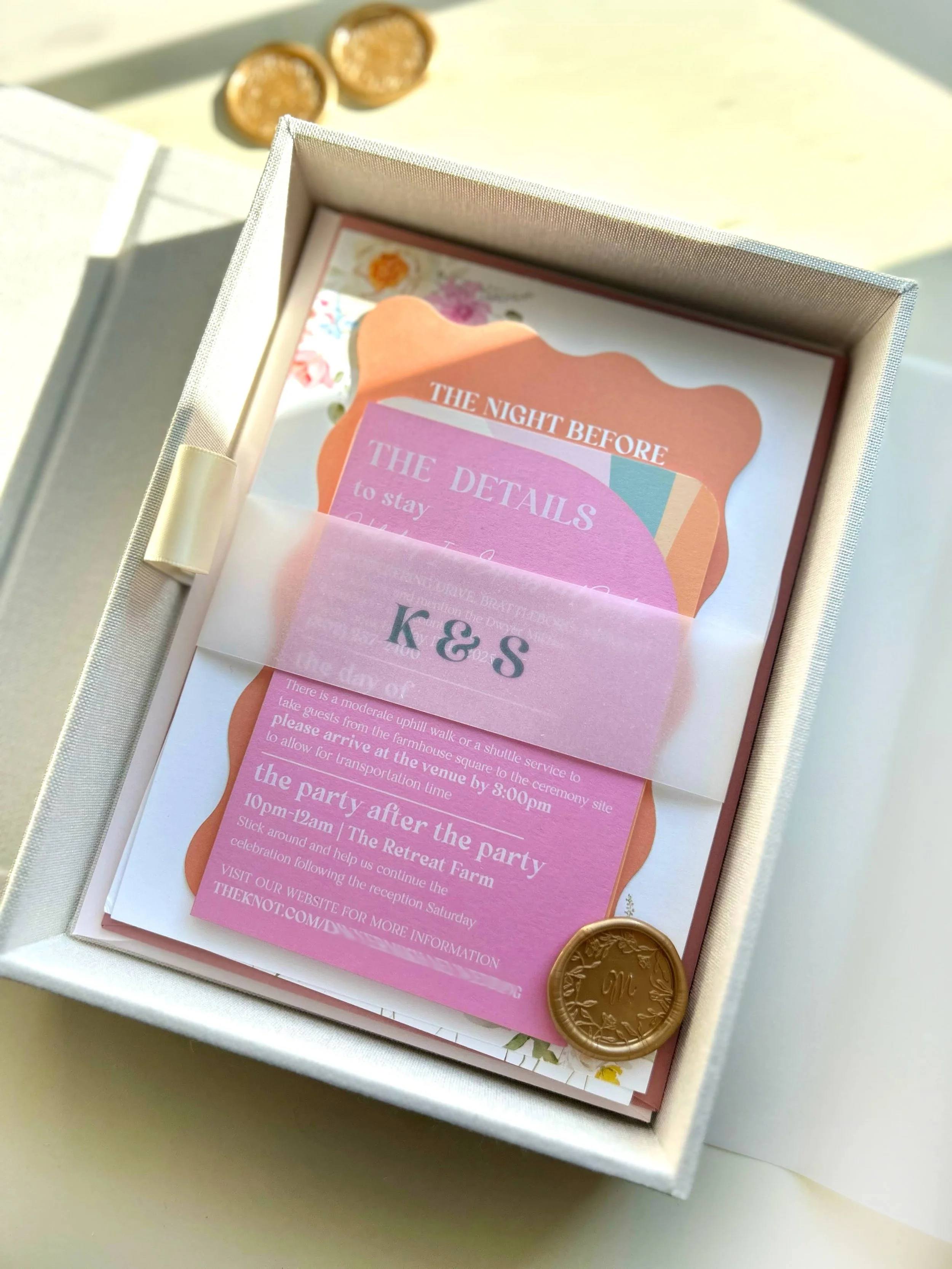 Wedding invitation inside a white box with a pink and orange design, gold wax seal, and a translucent pink band with the initials 'K & S' on it.