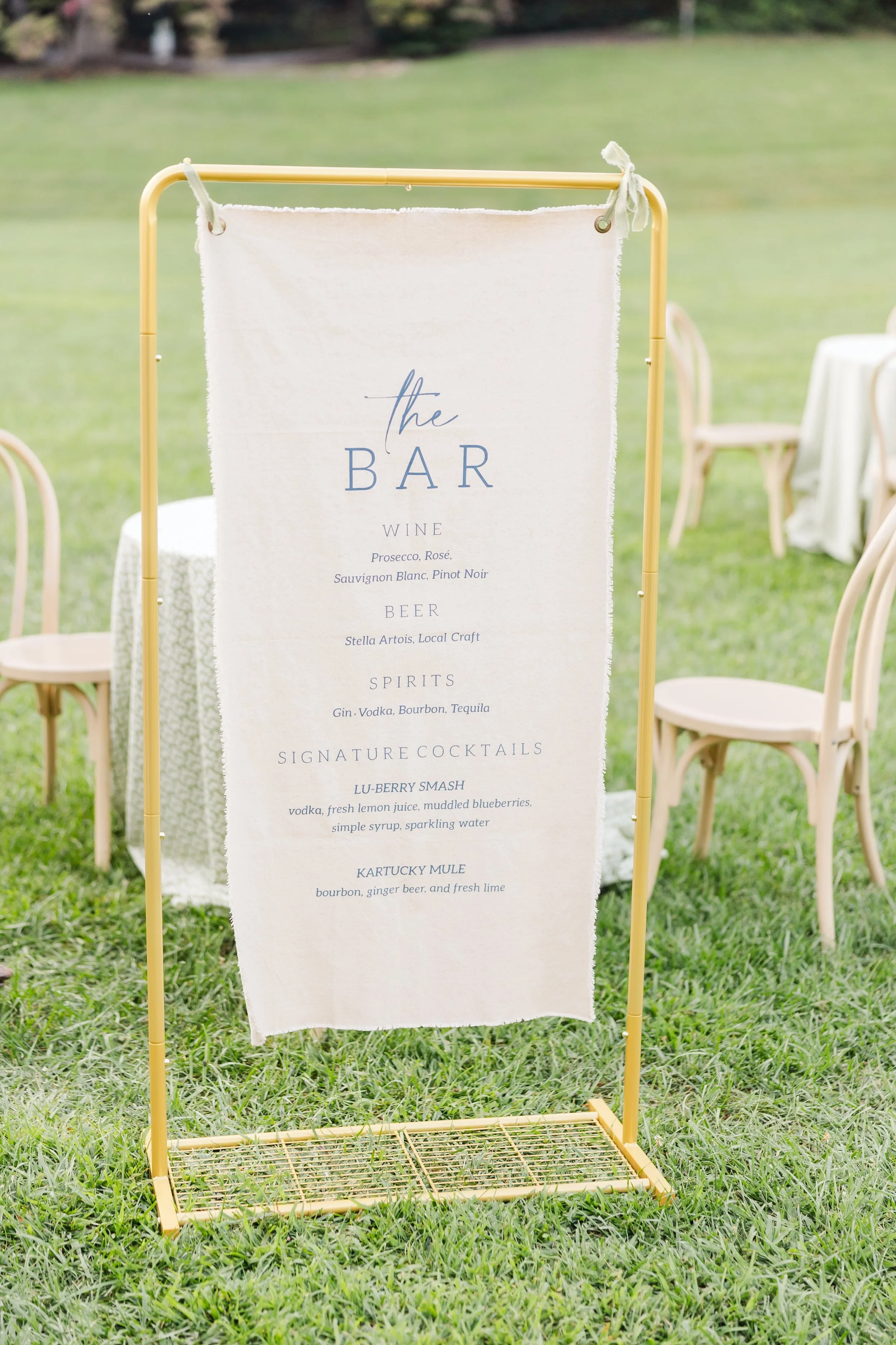 Wedding beverage menu on a white fabric banner with blue text, surrounded by light-colored chairs and green grass outside.