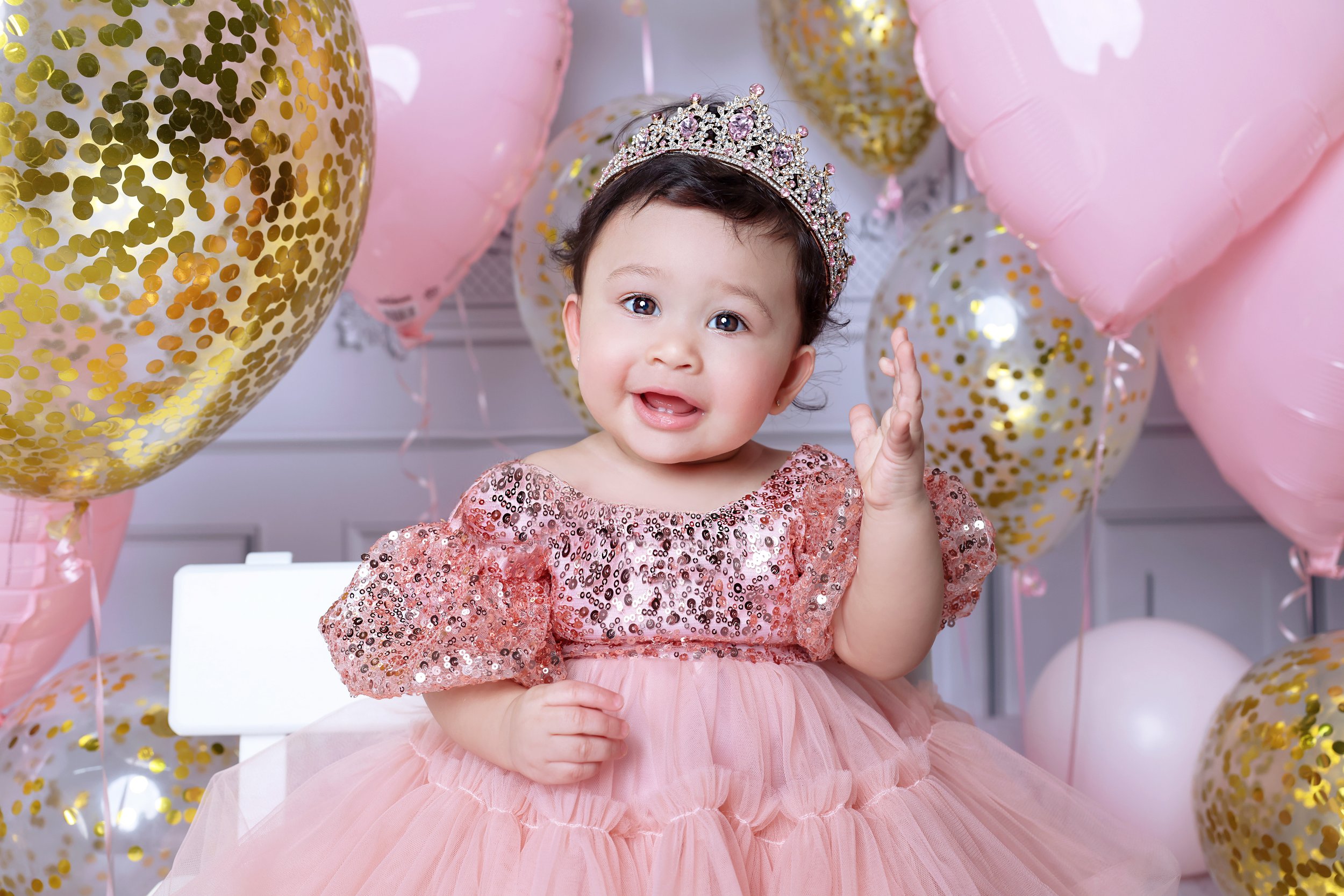 A young girl dressed in a pink sequined dress and wearing a tiara, celebrating her birthday surrounded by pink and gold balloons.