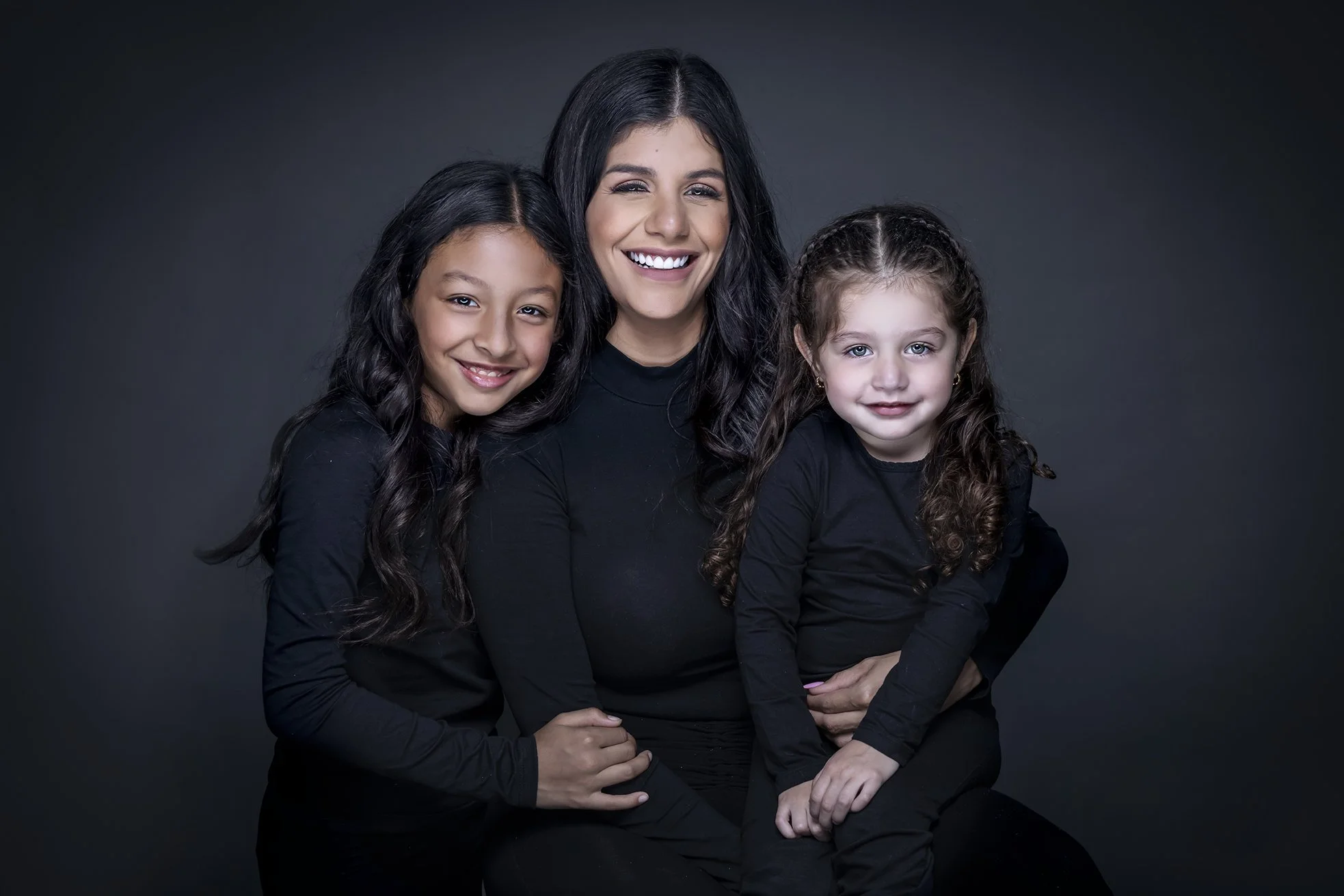 A woman with long dark hair smiling and winking, sitting with two young girls, all dressed in black, against a dark background.