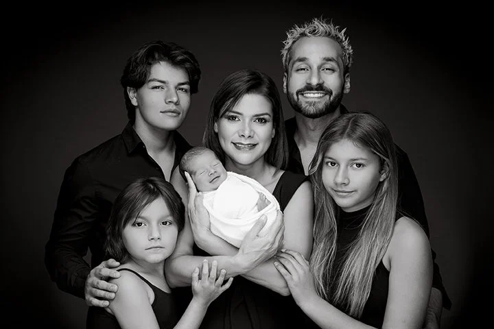 Black and white portrait of a smiling family with five members, including a newborn, standing close together against a dark background.