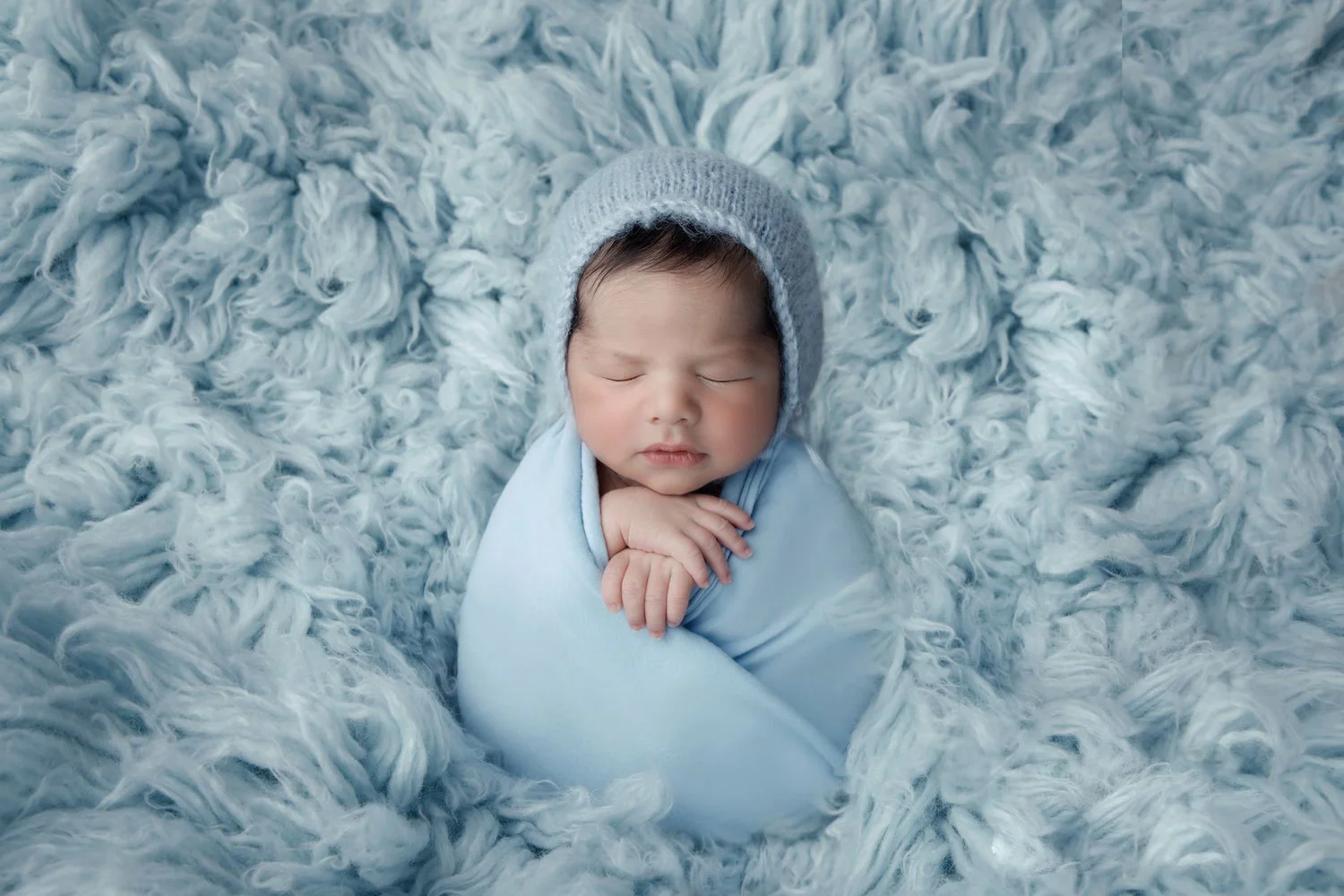 Close-up of a sleeping baby wrapped in a light blue blanket on a fluffy blue rug, wearing a gray knit cap.