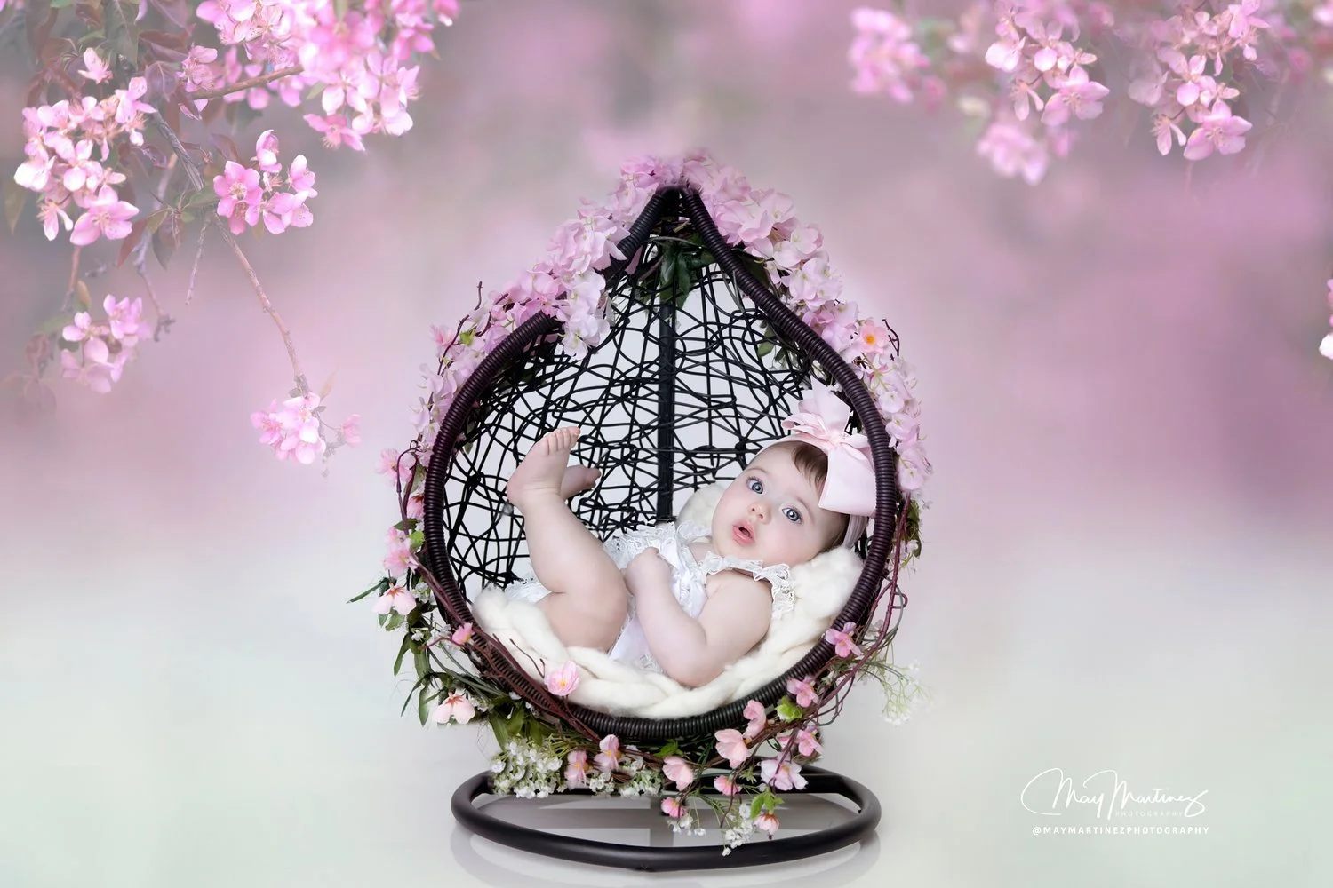 Baby girl lying in a decorative wicker swing surrounded by pink cherry blossoms.