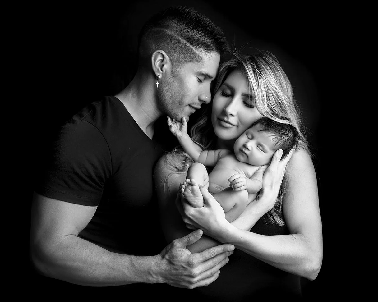 Black and white portrait of a man, woman, and baby. The man and woman hold the sleeping baby together, with the couple's faces close and eyes closed.