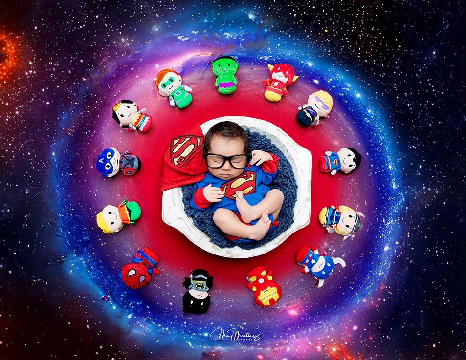 A child dressed as Superman sleeping on a white bed surrounded by plush toys of superheroes and cartoon characters against a cosmic galaxy background.