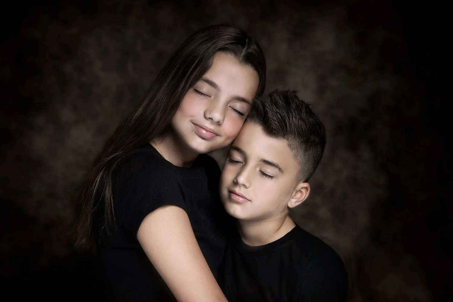 A young woman and a young boy with eyes closed embracing each other in a portrait against a dark background.