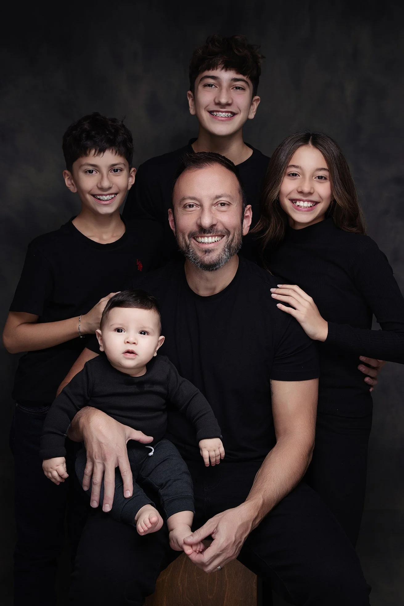 A happy family portrait with six members, including a man, a woman, and four children, all dressed in black, posing against a dark background.