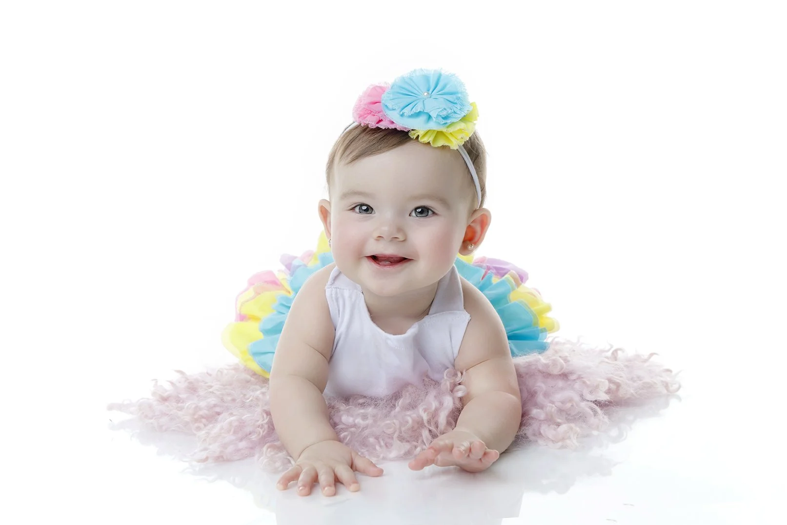 A baby girl with blue eyes, light brown hair, and a cheerful expression, lying on her stomach on a pink fluffy blanket, wearing a colorful tutu and a headband with pastel-colored fabric flowers.