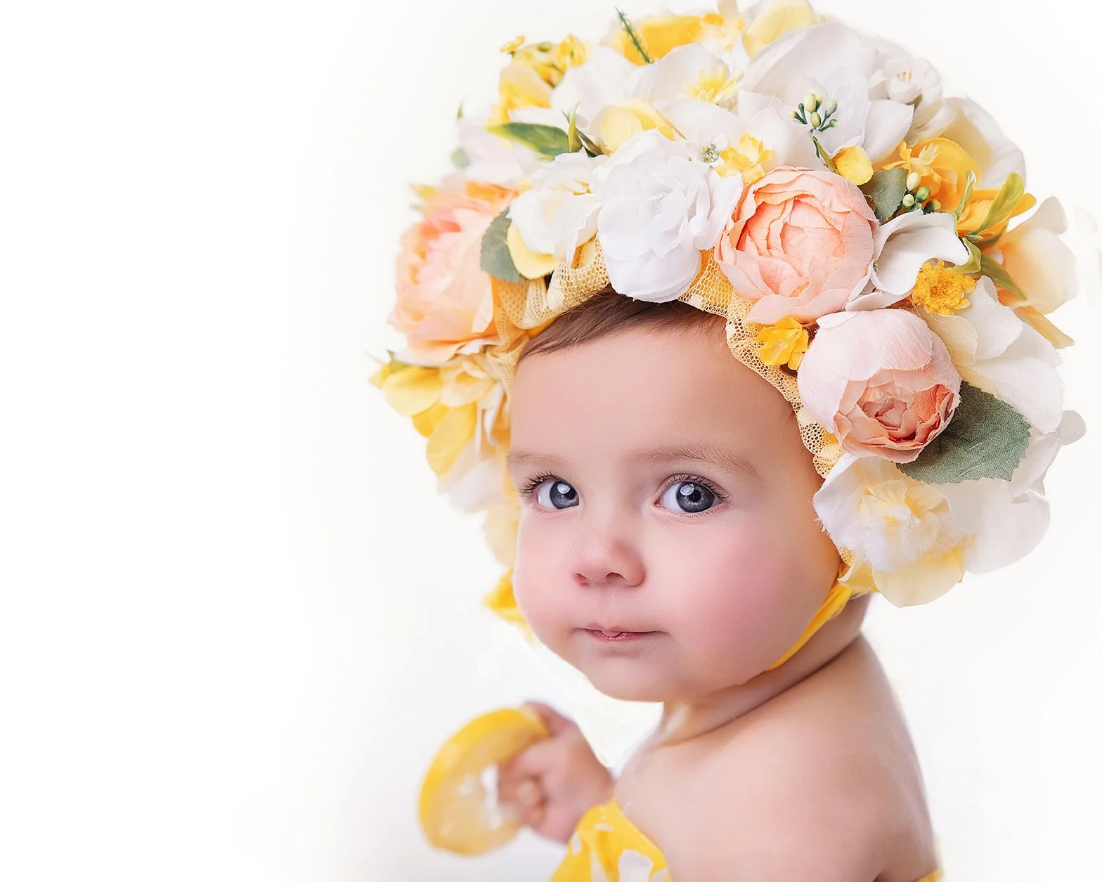 A baby girl wearing a large flower crown made of colorful flowers, holding a lemon slice, with a white background.